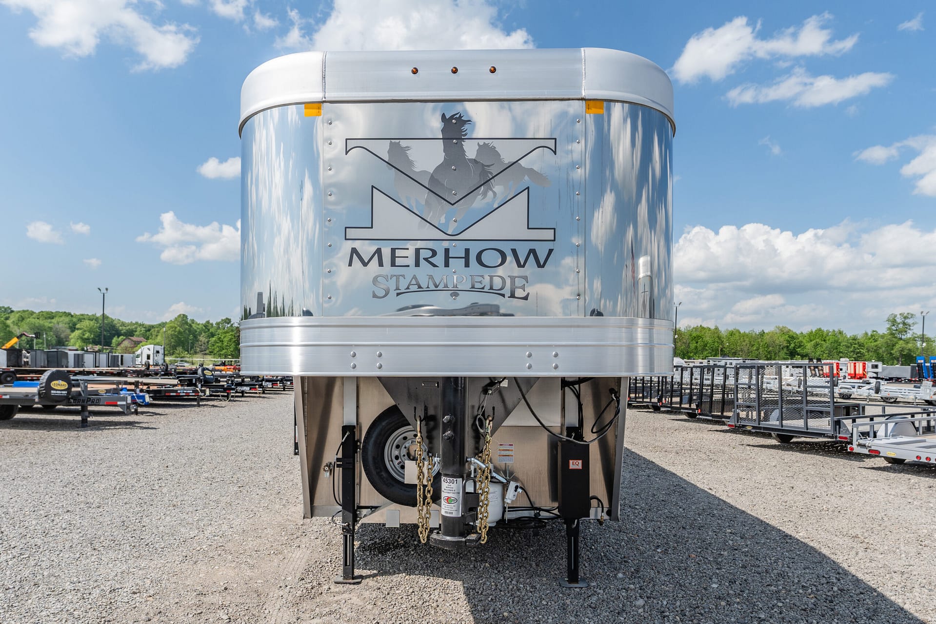Shiny aluminum horse trailer with a Merhow Stampede logo and horse silhouette on the front, parked in a lot under a blue sky.