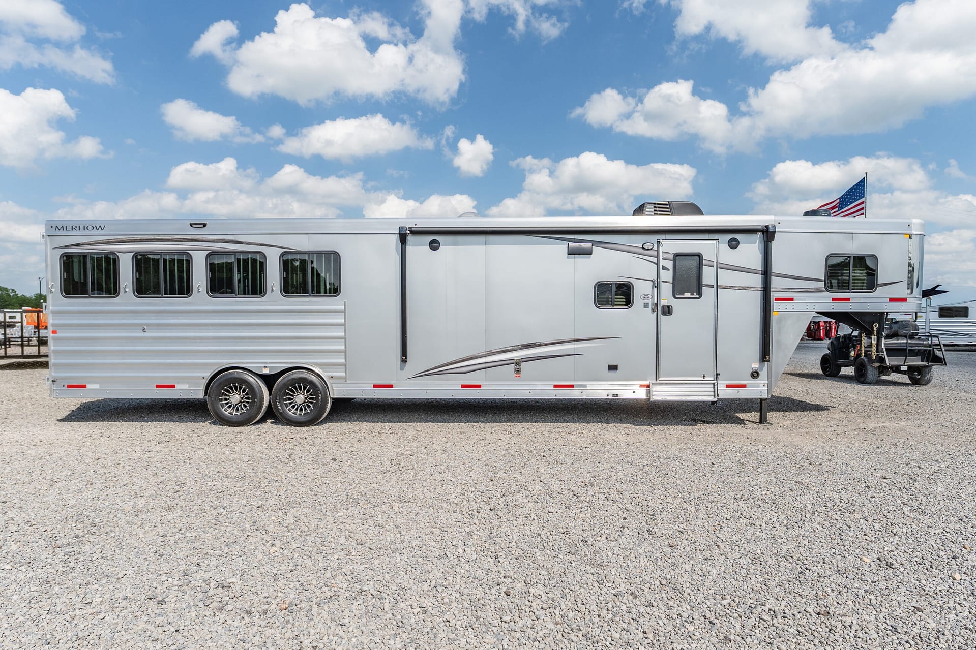 Side view of a silver Merhow horse trailer parked on a gravel lot under a blue sky with scattered clouds and an American flag visible at the rear.