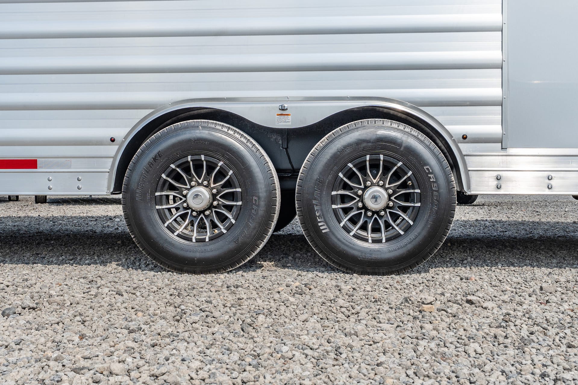 Side view of a silver trailer with two large black wheels on a gravel surface.