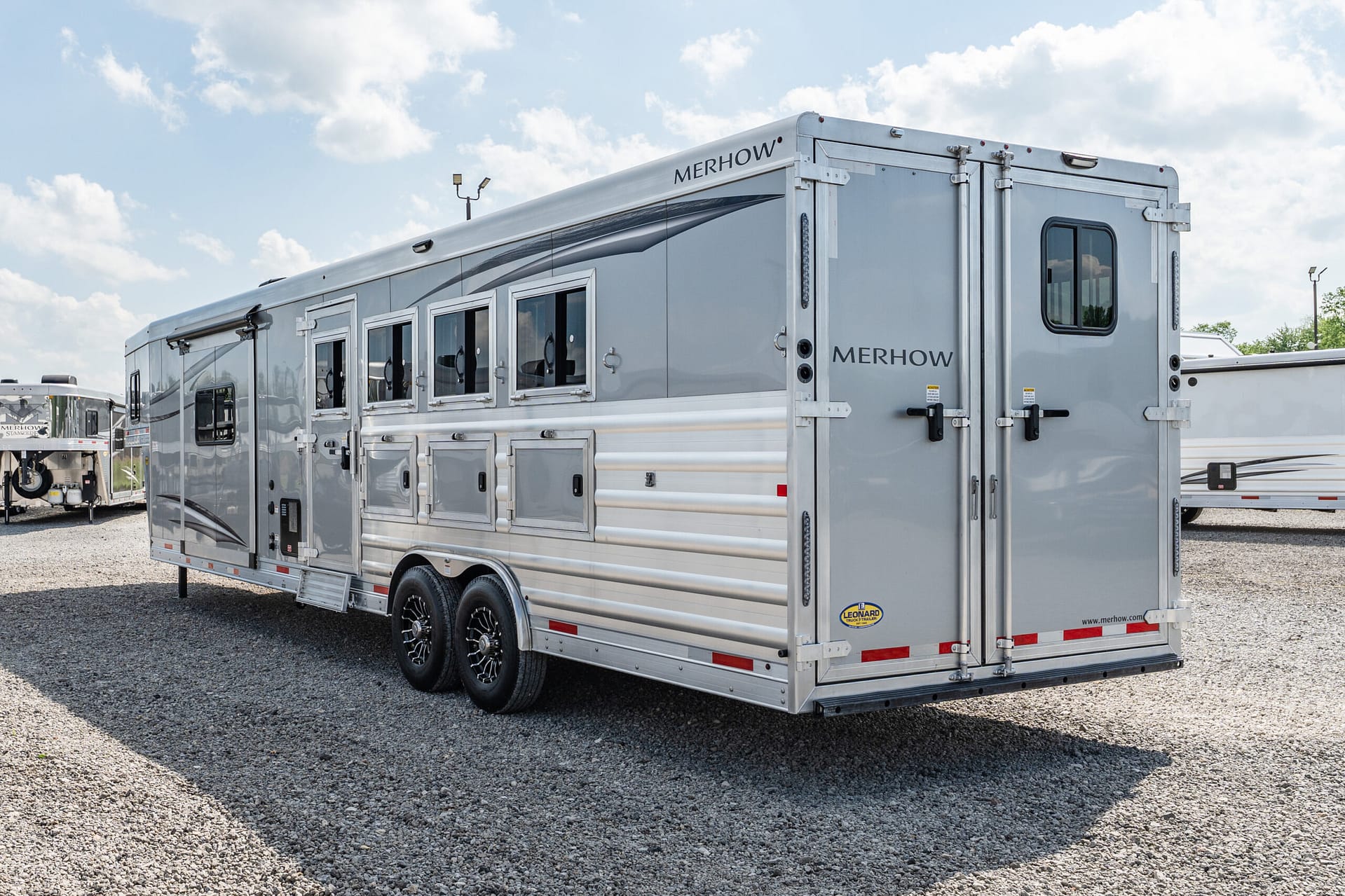 Silver Merhow horse trailer with multiple windows and rear double doors on a gravel lot.