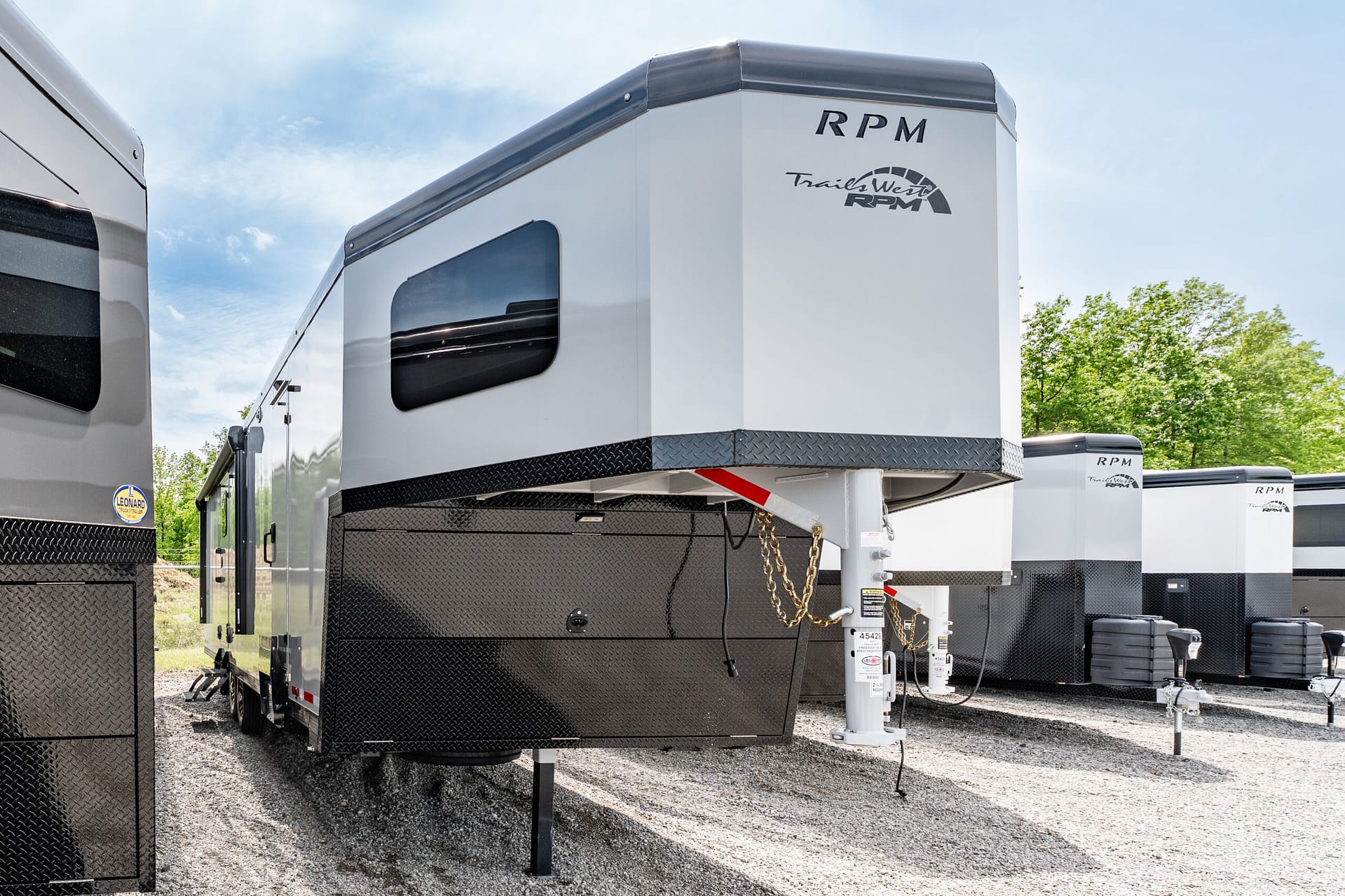 White RV trailer with black diamond-plate base and RPM Trail West logo, parked among other trailers on a gravel lot.