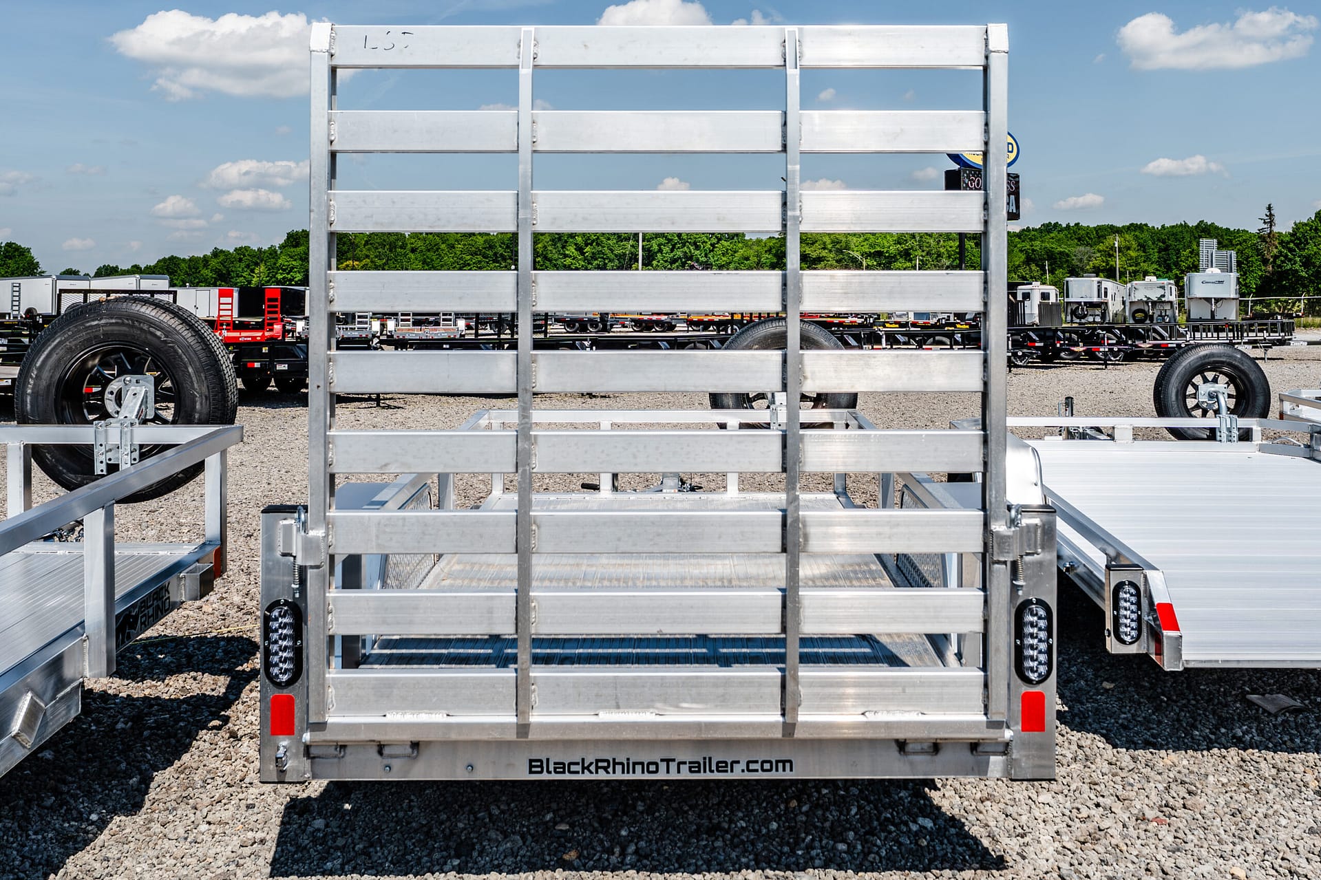 Rear view of a metal utility trailer frame on a gravel lot with other trailers in the background and 'BlackRhinoTrailer.com' on the bumper.