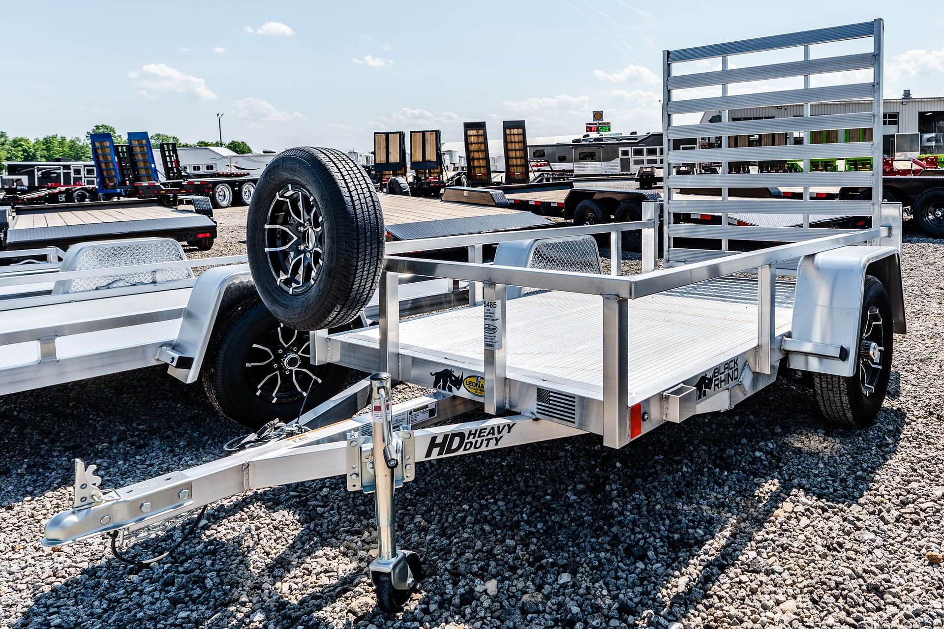 Front view of a flatbed trailer with a spare tire mounted on its frame, in a gravel lot with other trailers nearby on a sunny day