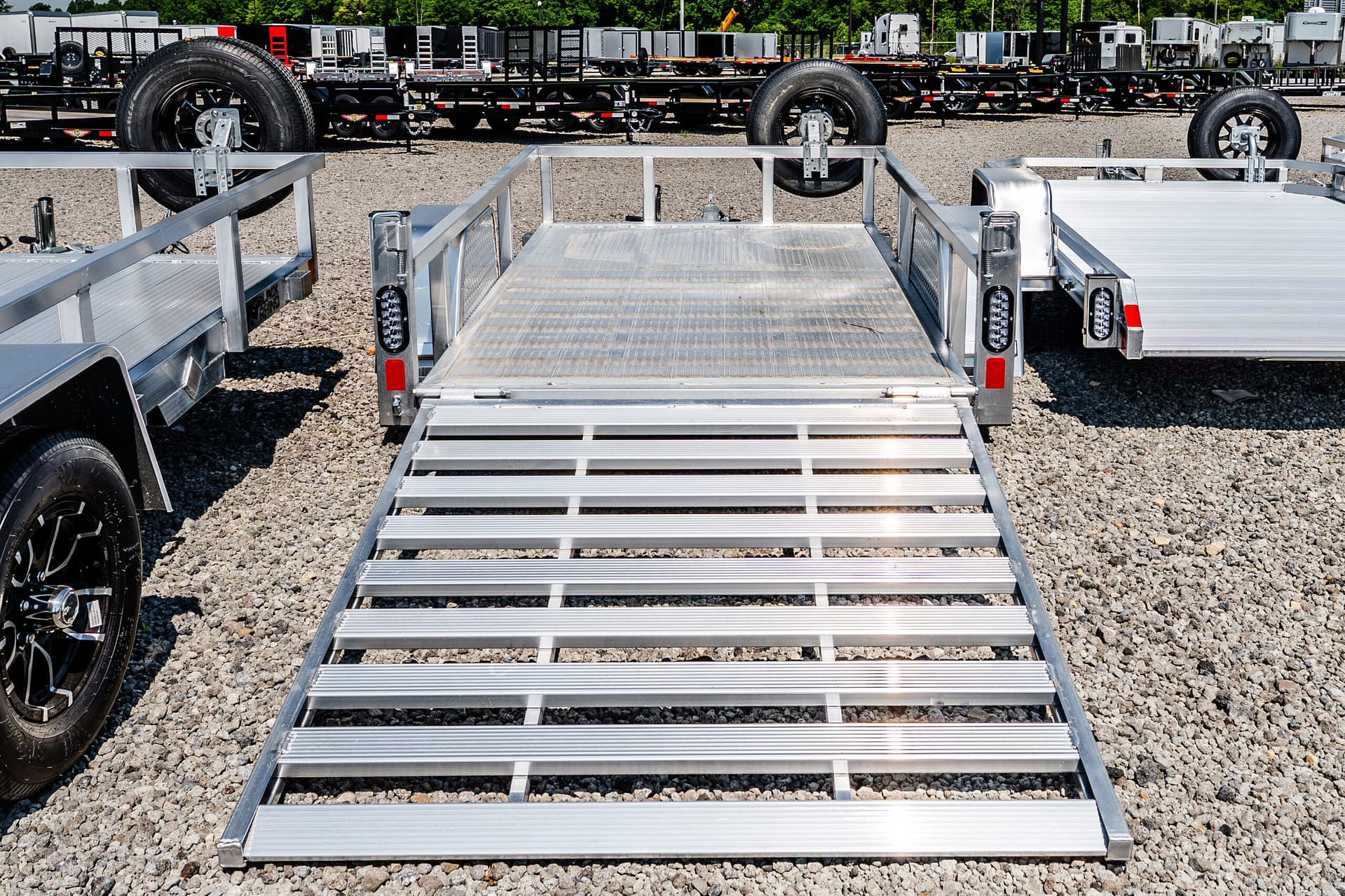 Wide shot of silver aluminum trailer ramps extending toward the camera in a gravel lot, with numerous trailers and equipment in the background.