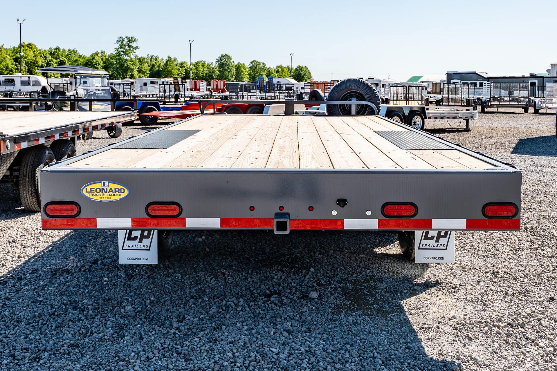Rear view of a flatbed trailer in a gravel lot with other trailers and a Leonard Truck & Trailer logo visible on the side plate.