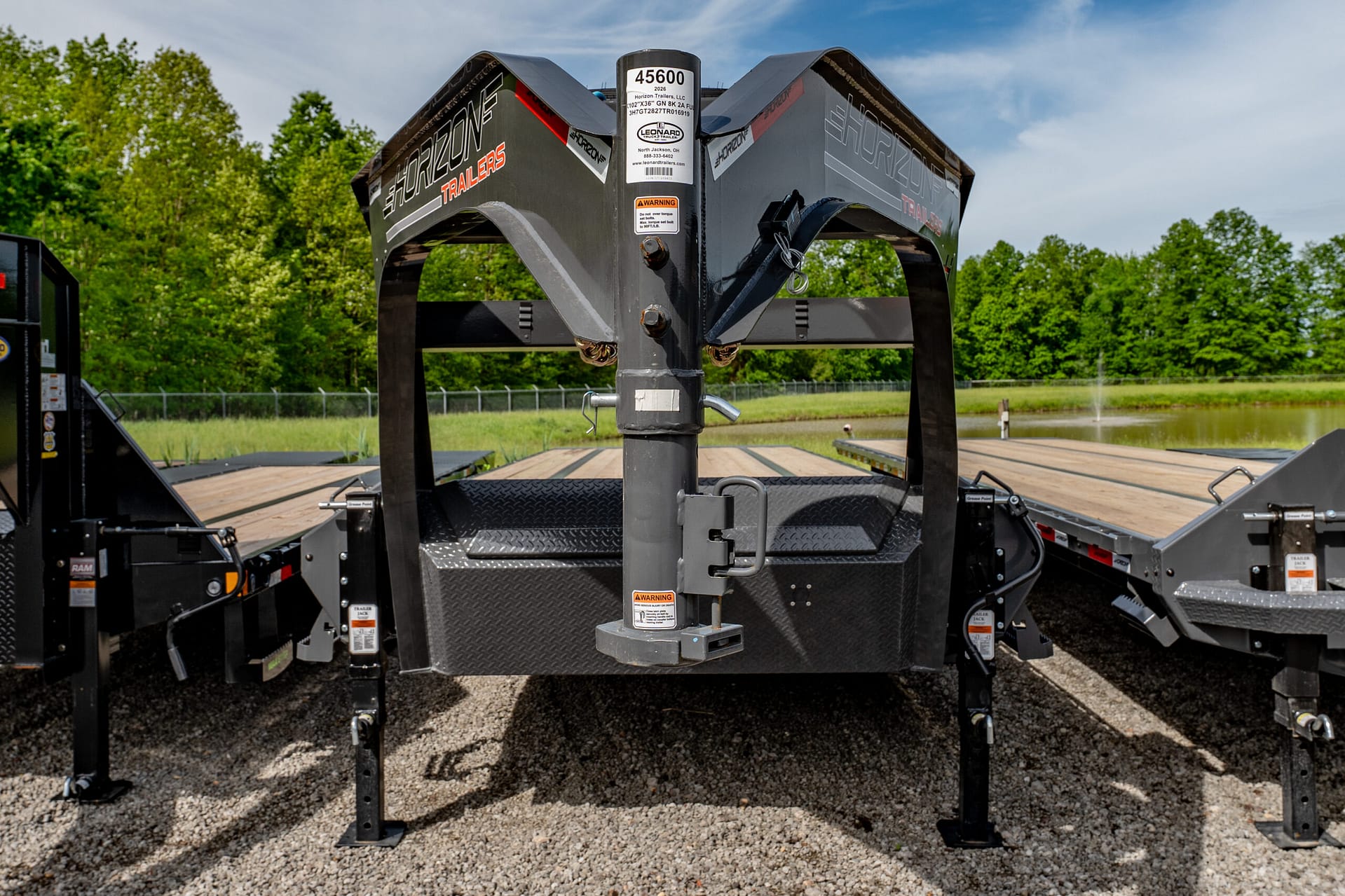 Front view of a heavy-duty trailer hitch and frame on a gravel lot, with green trees in the background.
