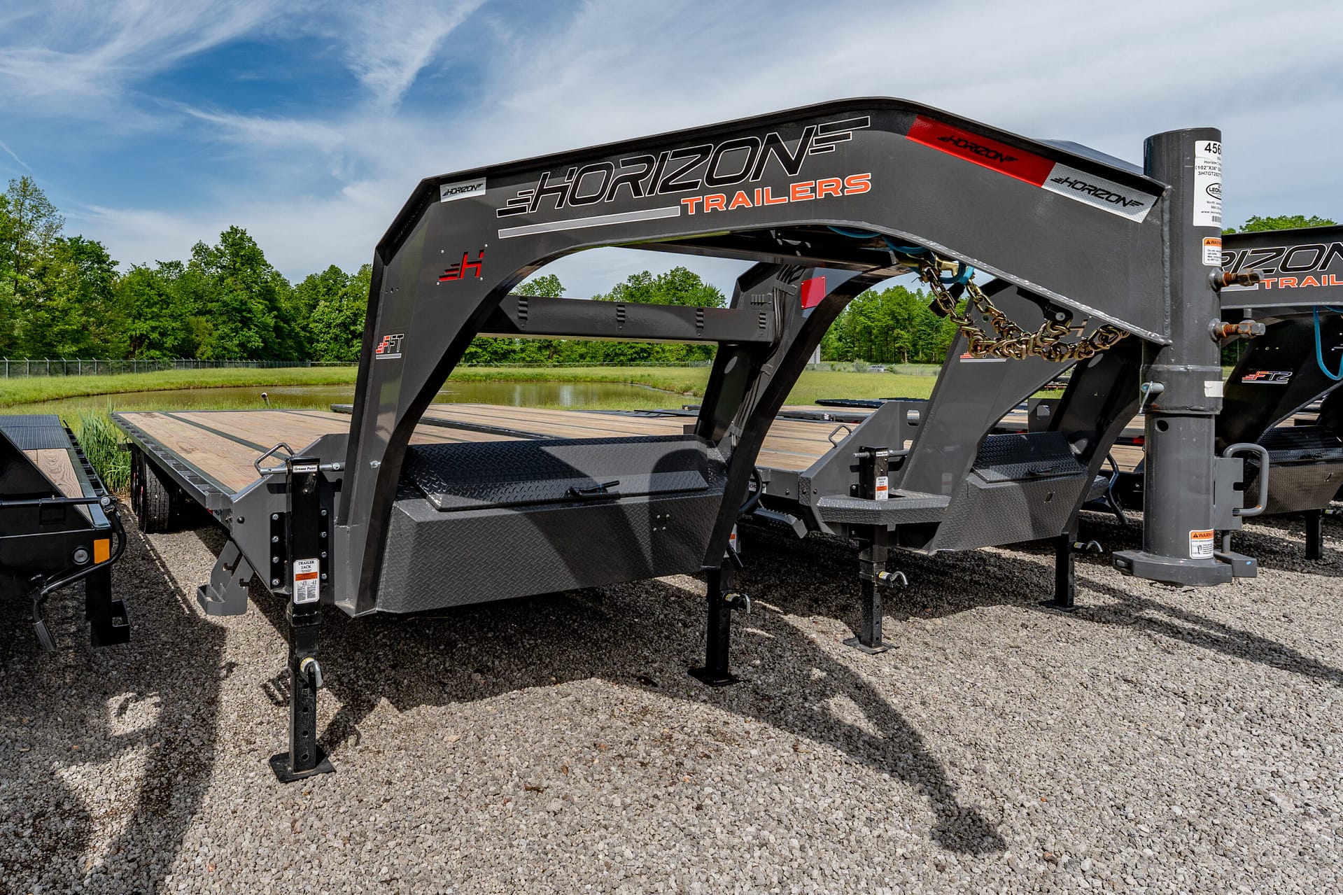 Large Horizon Trailers gooseneck hitch frame with chains, mounted on a gravel lot under a blue sky and green trees in the background.