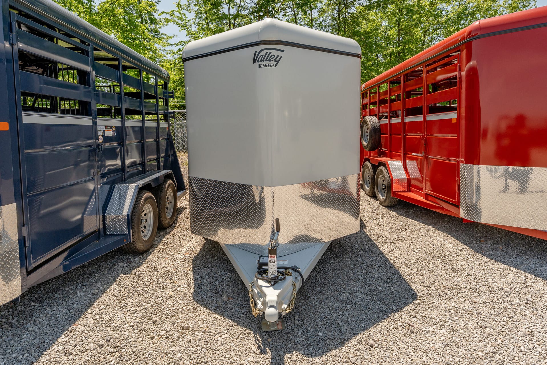 Gray enclosed trailer between blue and red trailers on a gravel lot, with a hitch and chains in the foreground.