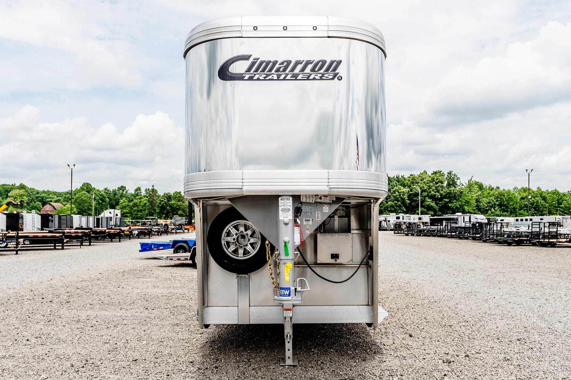 Shiny silver semi-trailer front curb with a large spare tire visible in the open bay and a bright, cloudy sky behind.