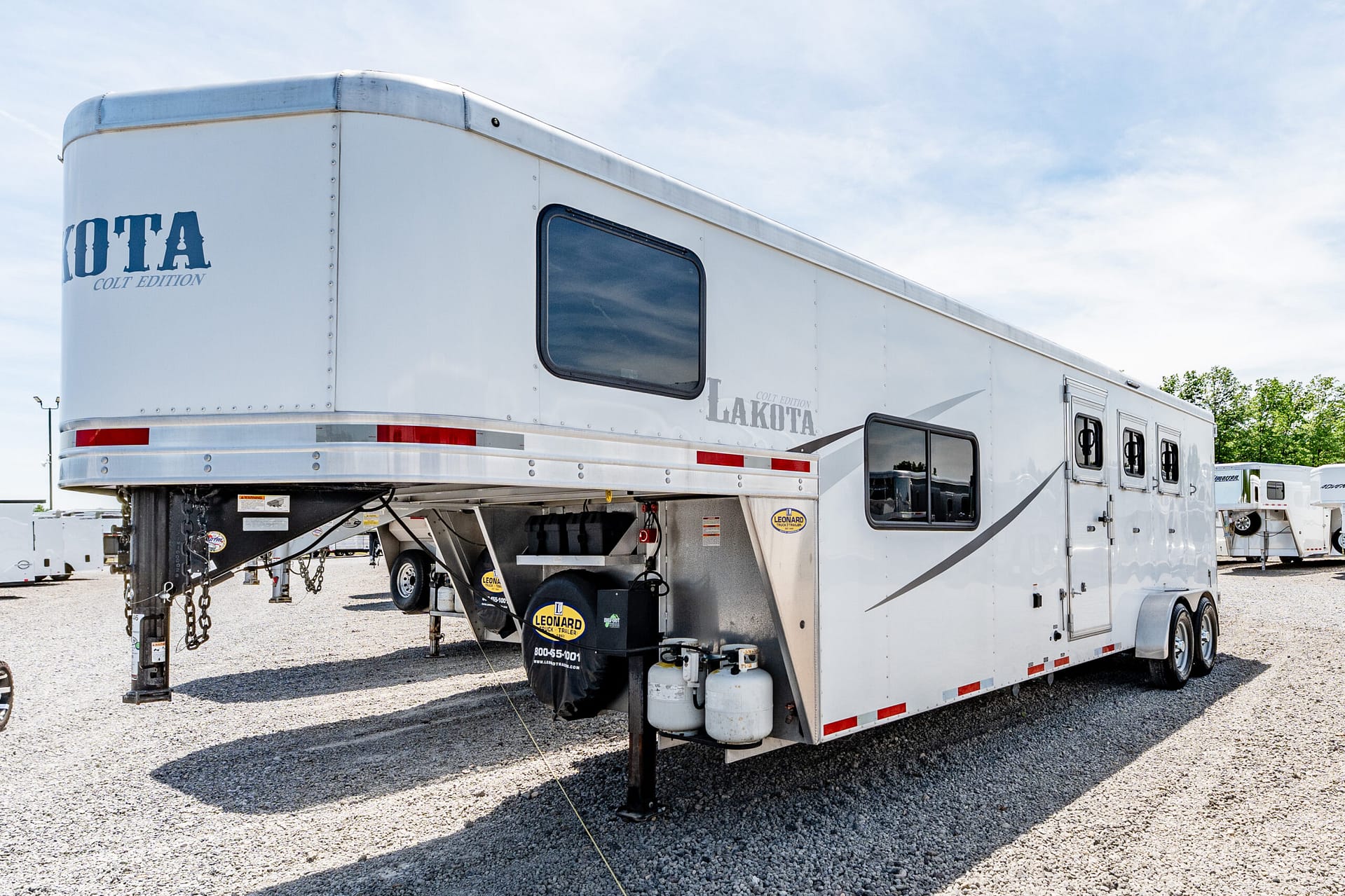 White Lakota Edition gooseneck horse trailer viewed from the side and rear on a gravel lot under a blue sky with windows visible on the side.