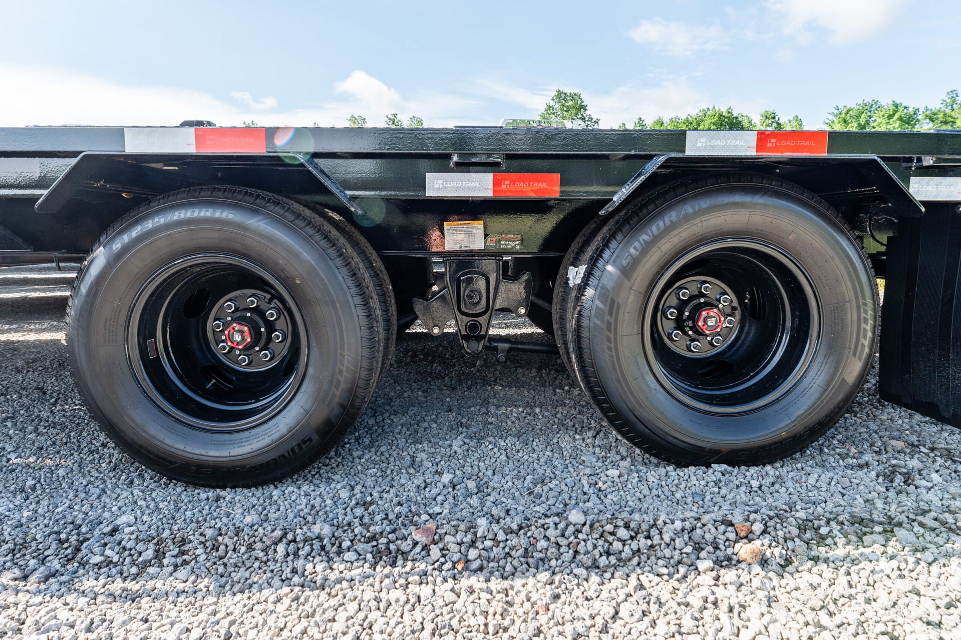 Dual-axle trailer wheels with black rims and red center caps on a gravel surface near a flatbed trailer.",