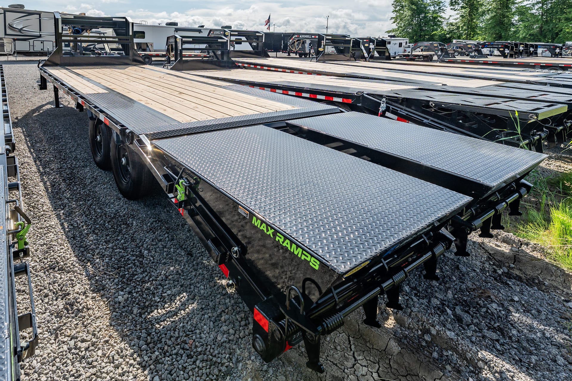 Row of black flatbed trailers with wooden decks and diamond-plate ramps parked in a gravel lot.