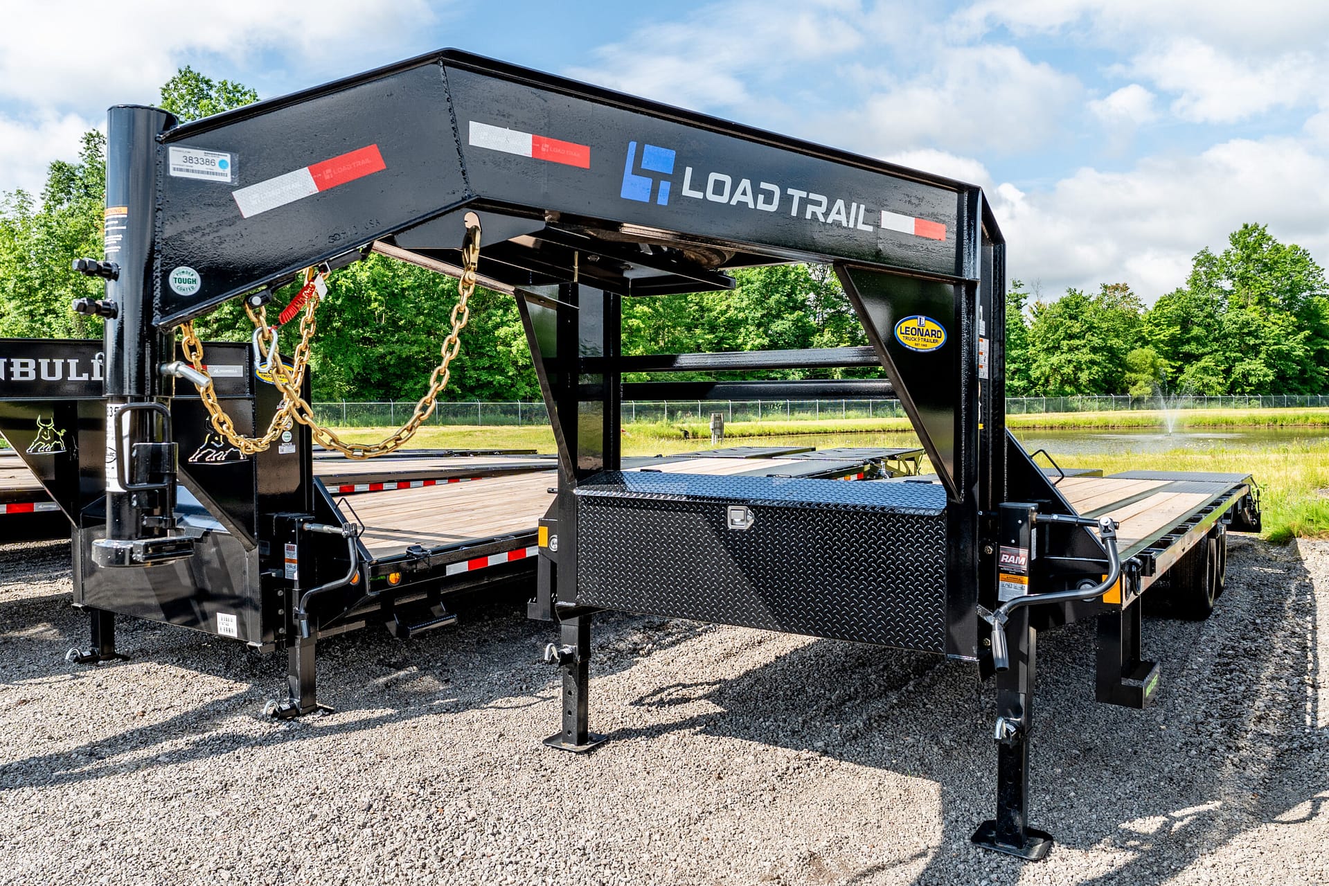Wide view of a heavy-duty black gooseneck trailer frame with chains, decals, and a diamond-plate toolbox, parked on gravel with trees in the background.