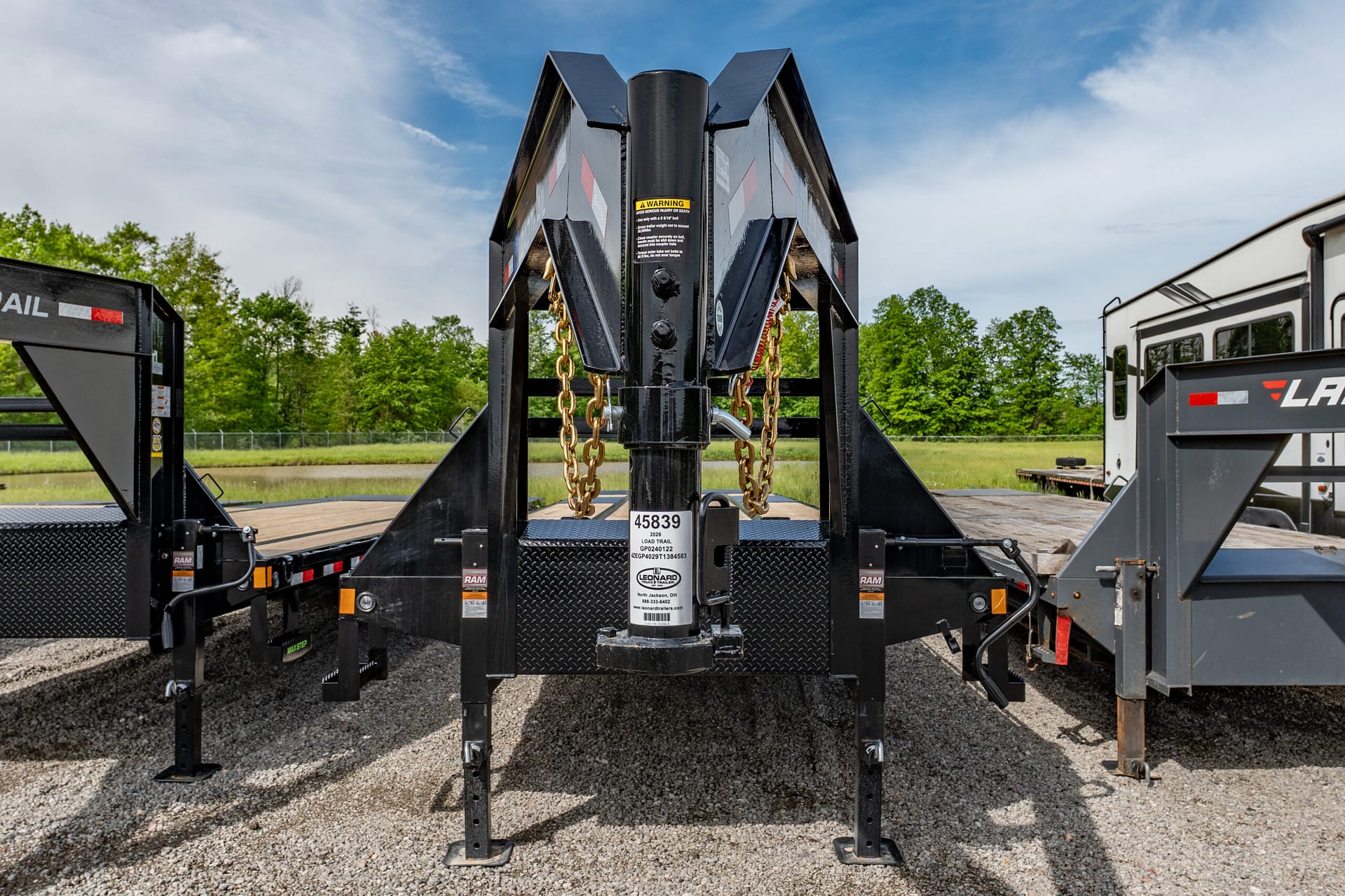 Center-front view of a heavy-duty trailer hitch with chains and warning labels, on a gravel lot with trees and a blue sky behind.