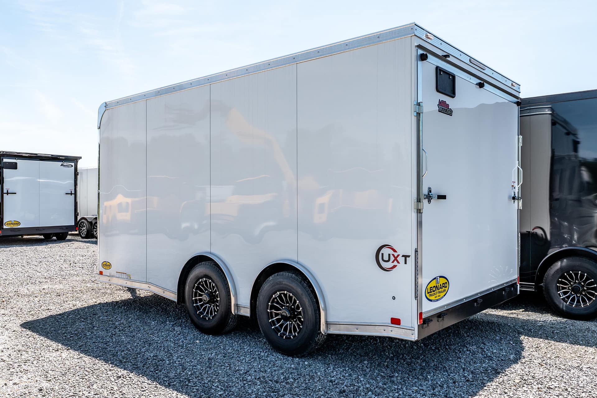 White enclosed cargo trailer with triple axles and alloy wheels parked on a gravel lot; branding stickers visible along the side edge.