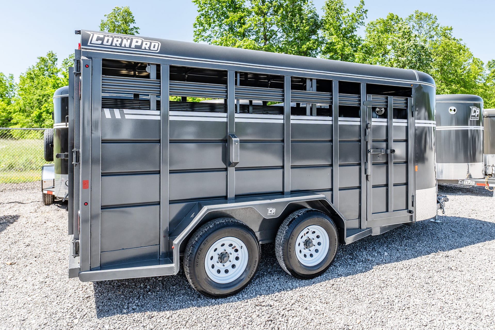 Dark gray livestock trailer with barred side vents and dual rear doors, parked on gravel with green trees in the background, brand name visible on the front edge.