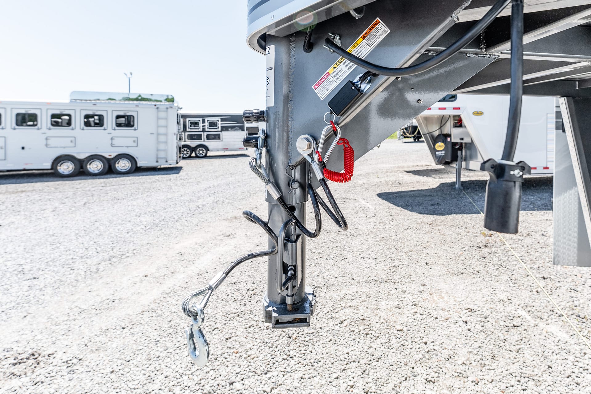 Close-up of a trailer hitch with safety chains, cables, and labels, in a gravel lot with horse trailers in the background.