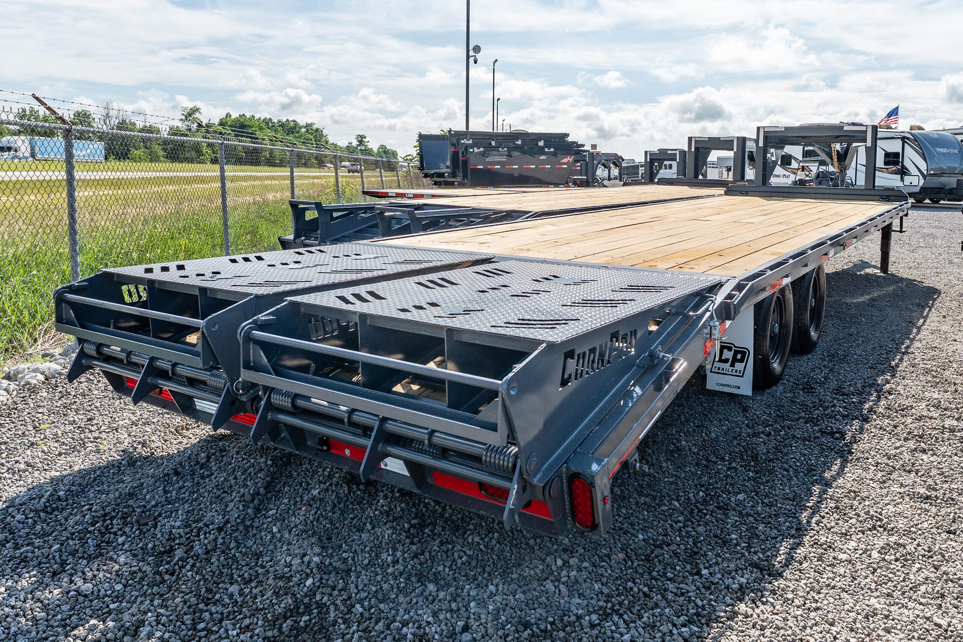 Wide view of a heavy-duty flatbed trailer with wooden deck and metal ramps on a gravel lot, fence in foreground and other trailers in background, under a blue sky with clouds.