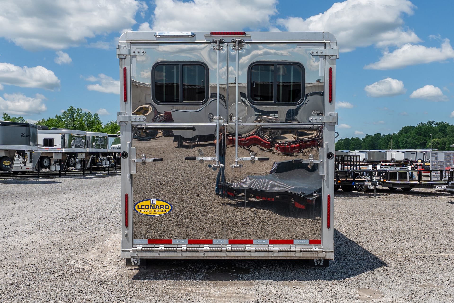 Rear view of a polished silver trailer with dual doors, red lights, and a Leonard Truck & Trailer logo at the bottom left amid a gravel lot and blue sky.