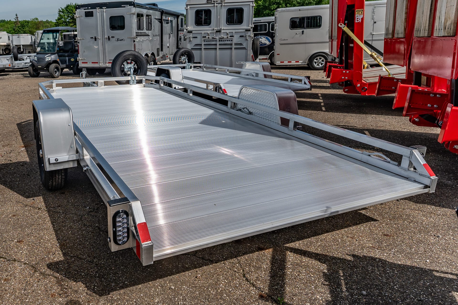 Aluminum flatbed utility trailer in a yard with horse trailers and red equipment in the background, viewed from the side.”,