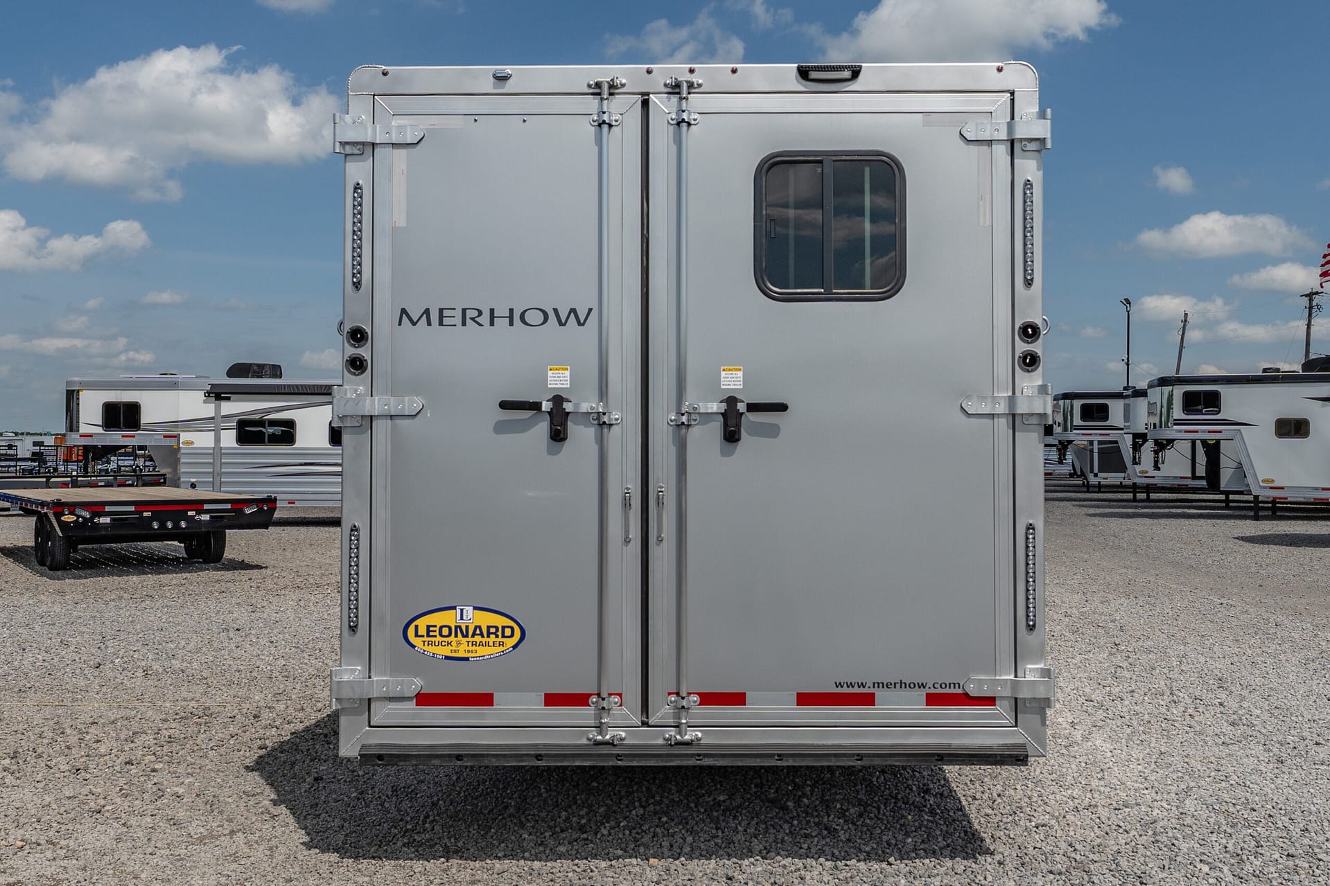 Rear of a silver Merhow horse trailer with two doors, a small window, and a Leonard trailer sticker on a gravel lot under a blue sky.