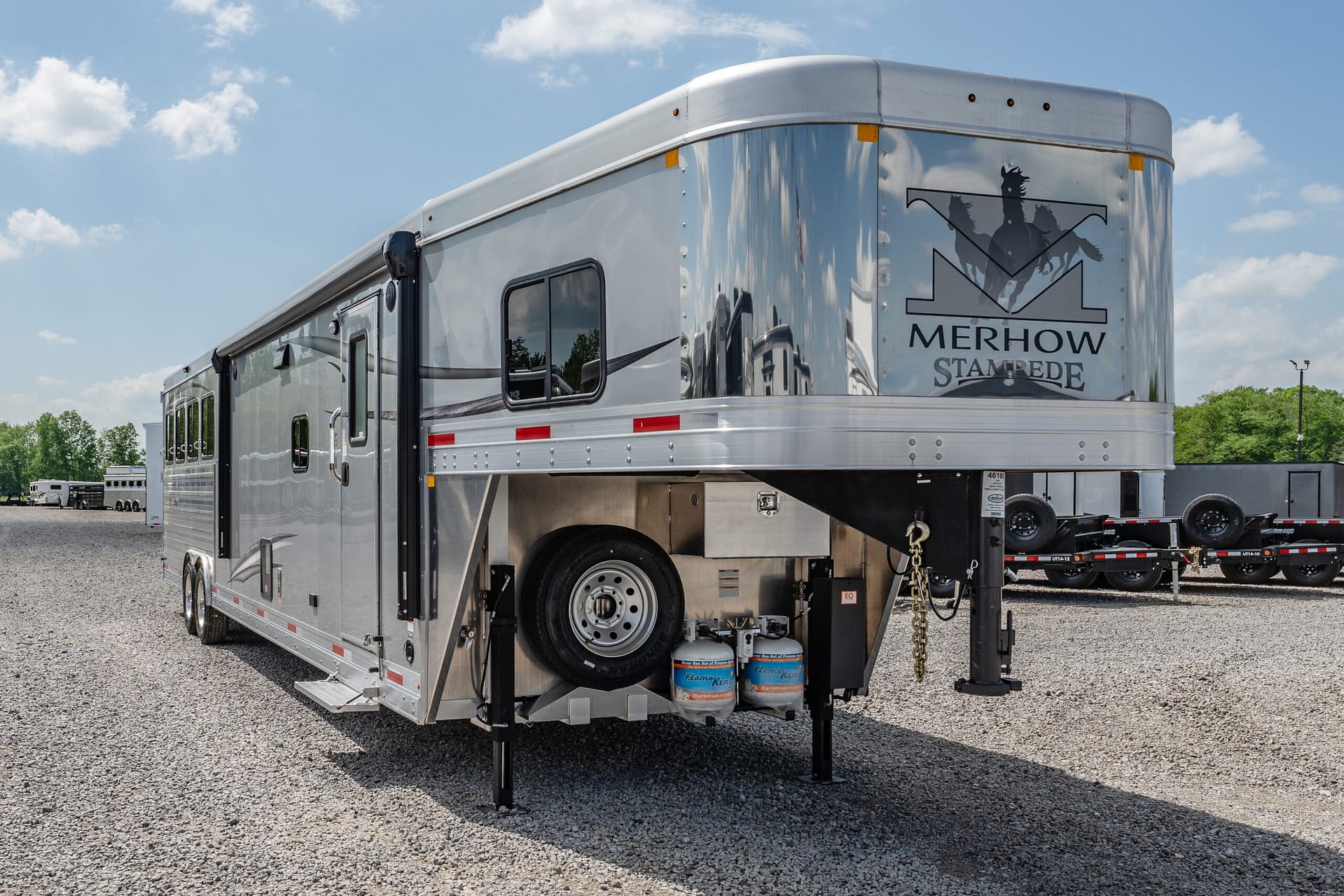 Wide view of a large silver Merhow Stampede horse trailer parked on a gravel lot under a blue sky with scattered clouds, logo visible on rear corner.