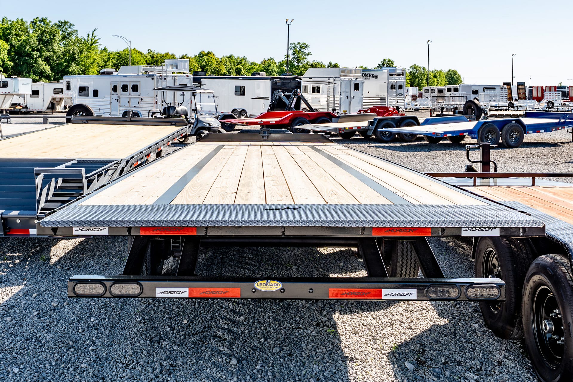 Row of white horse trailers and flatbed trailers parked in a gravel lot, with a wooden trailer deck in the foreground and green trees in the background.