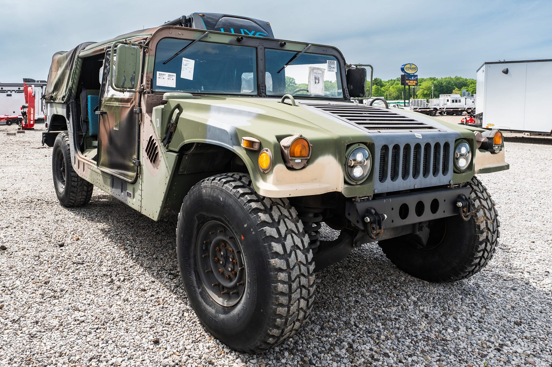 Military green off-road jeep with large tires parked on a gravel lot; other trailers visible in the background