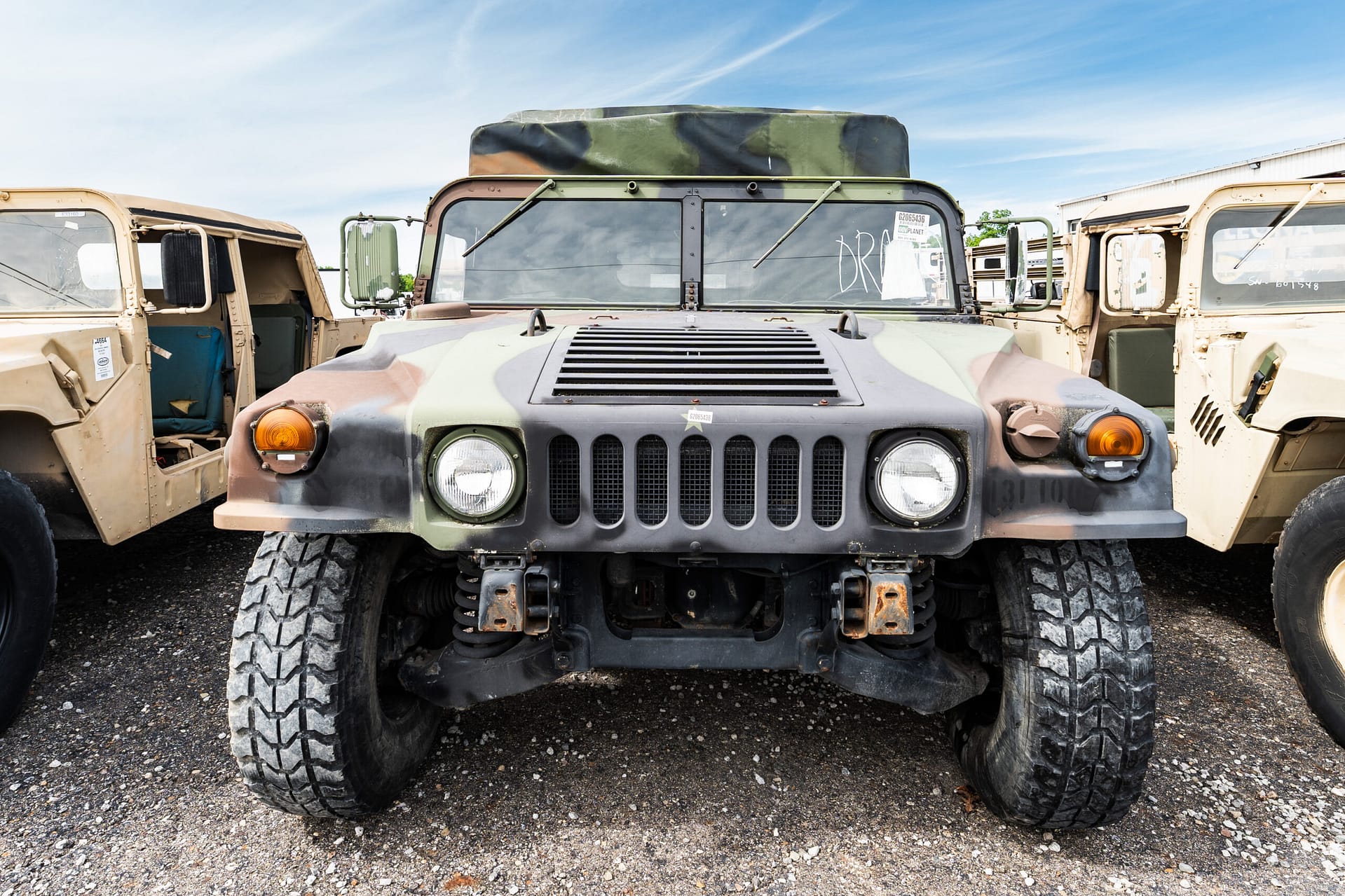 Front view of a camouflage military jeep with large tires, flanked by beige military vehicles with open doors under a blue sky.