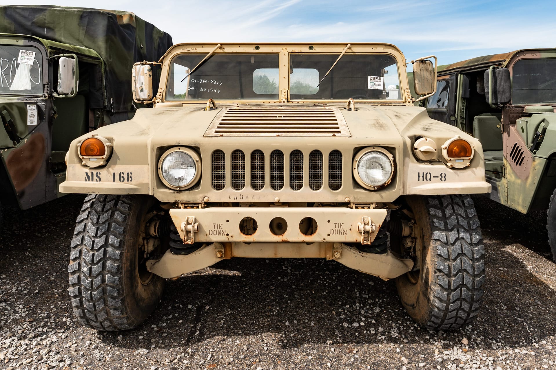 Front view of a weathered beige military Humvee with large off-road tires, flanked by other armored trucks on a gravel lot under a blue sky.