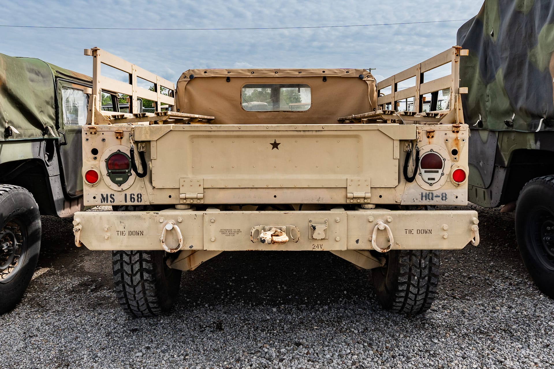 Rear view of a beige military cargo truck between two camouflaged trucks on a gravel lot, showing a small star insignia and rugged tires.