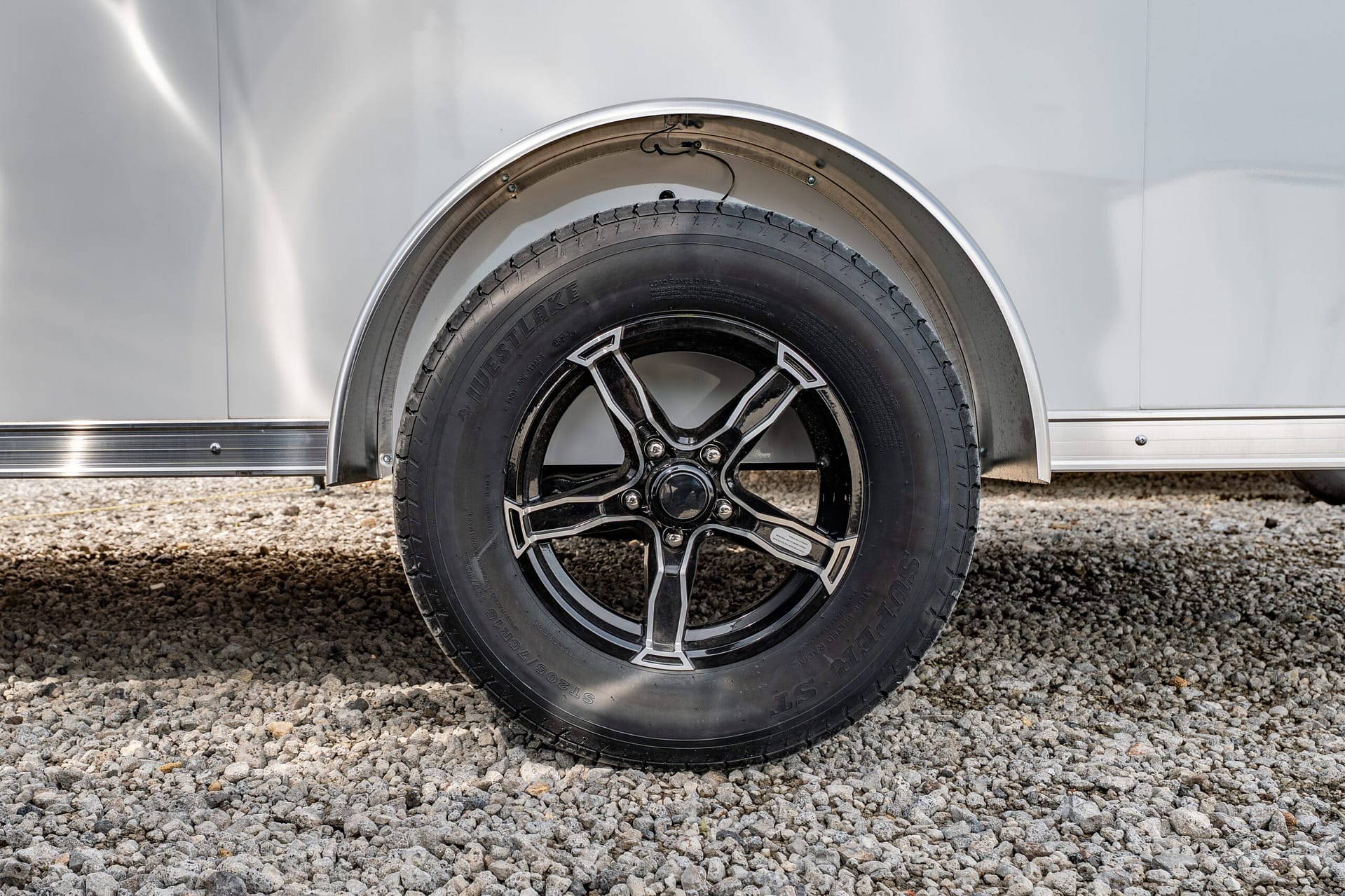 Close-up of a black alloy trailer wheel and tire inside a curved silver fender on a gravel surface, ground visible beneath.