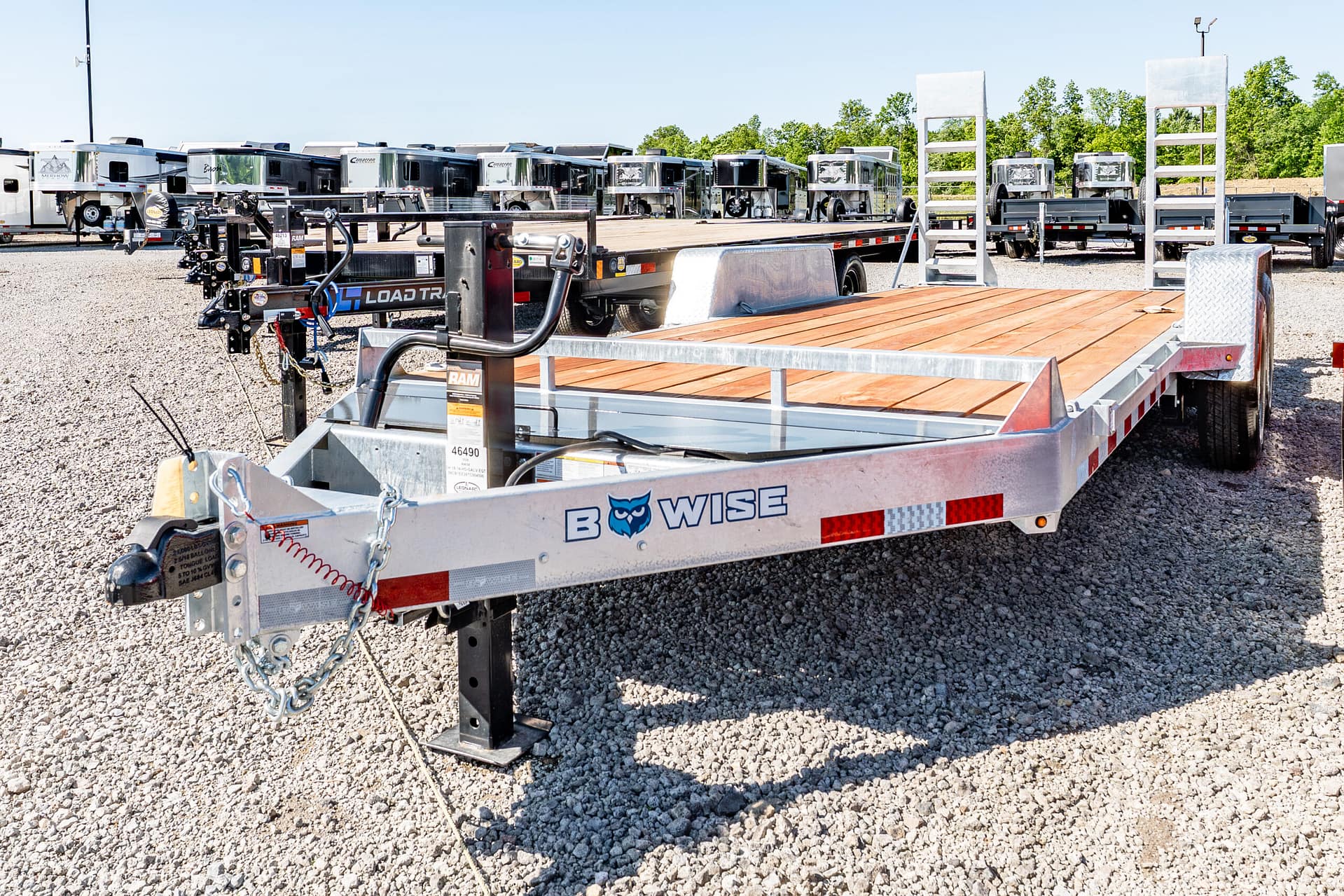 foreground: a flatbed trailer with a wooden bed and metal frame in a gravel lot, with towing chains and equipment visible; row of RVs and trailers in the background.