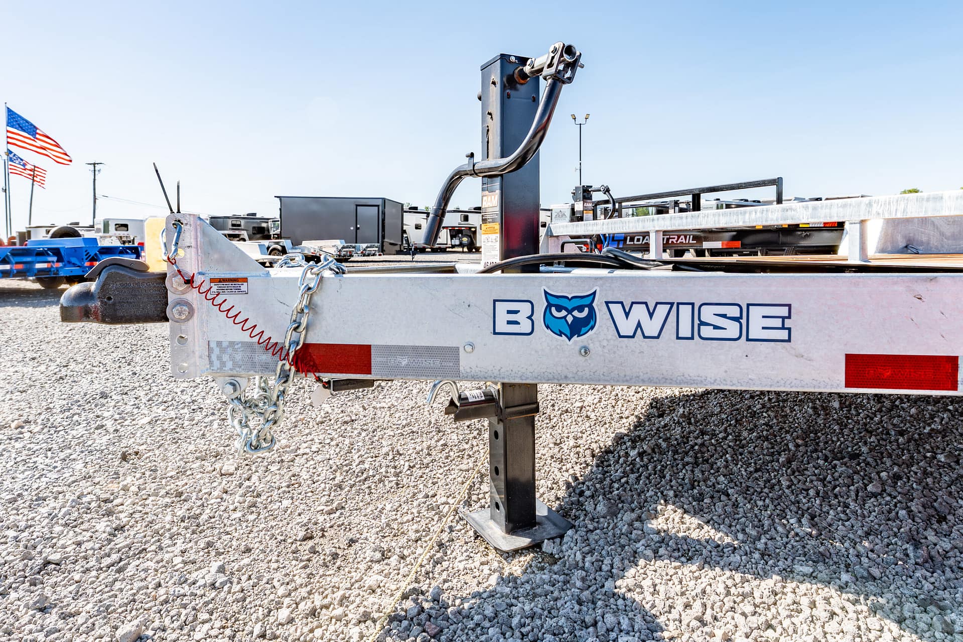 Close-up of a trailer frame with a hitch and safety chains on gravel, with flags in the background and clear blue sky.