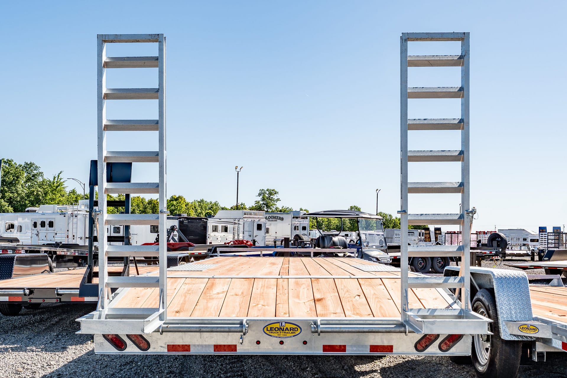 Rear view of a flatbed trailer with tall metal ladder frames on both sides in a truck yard, with other trailers in the background.