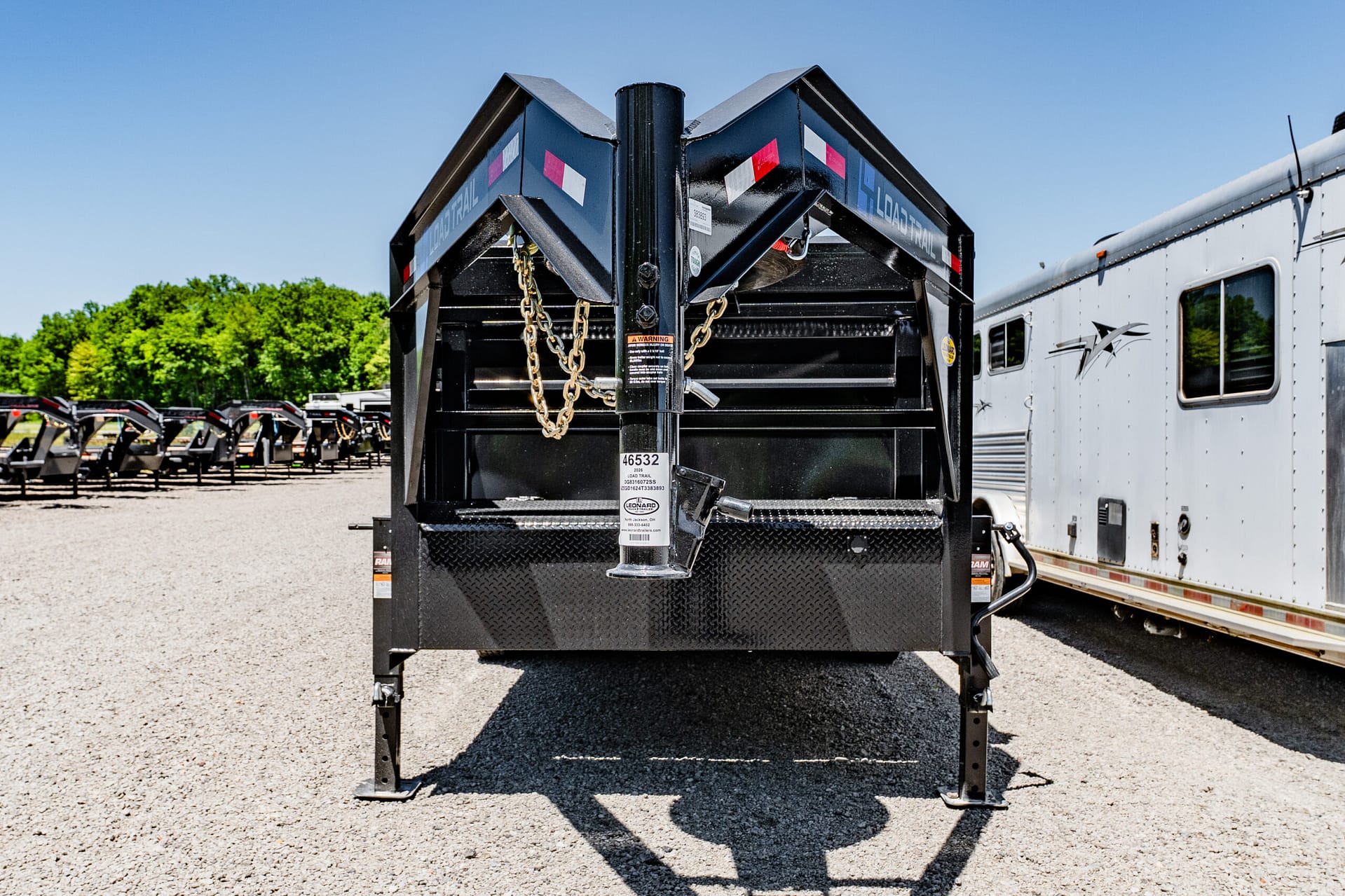 Rear view of a black dump trailer with a V-shaped hitch, safety chains, and warning stickers, parked in a lot beside a white trailer to the right.