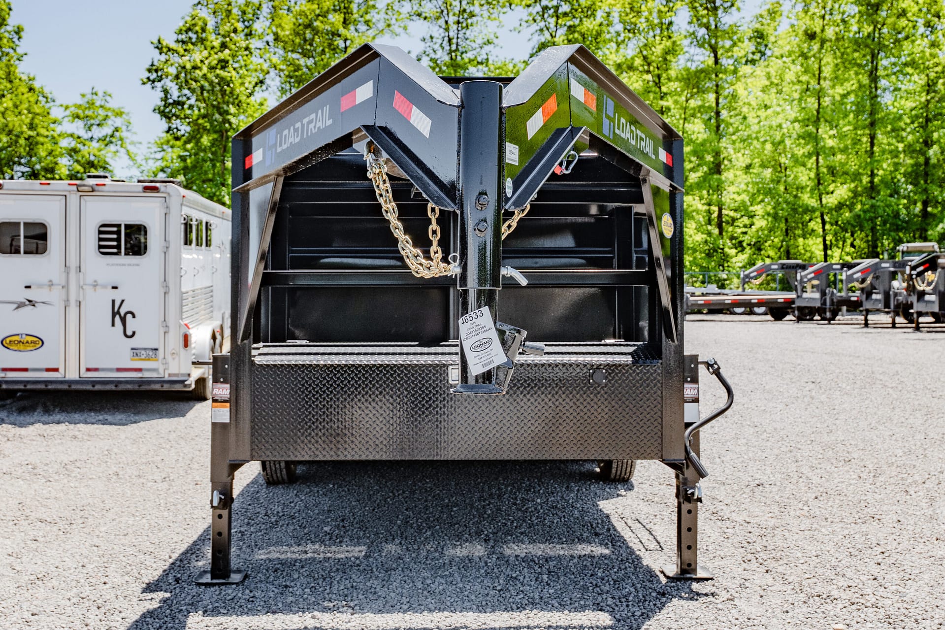 Front view of a black dump trailer with safety chains and hitch, in a gravel lot with trees and other trailers in the background.
