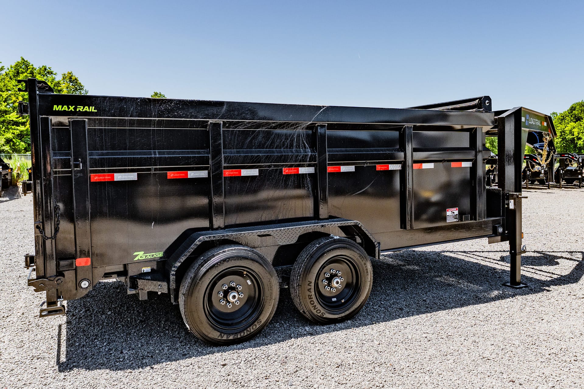 Side view of a large black Max Rail dump trailer with dual axles and reflective strips, parked on gravel.