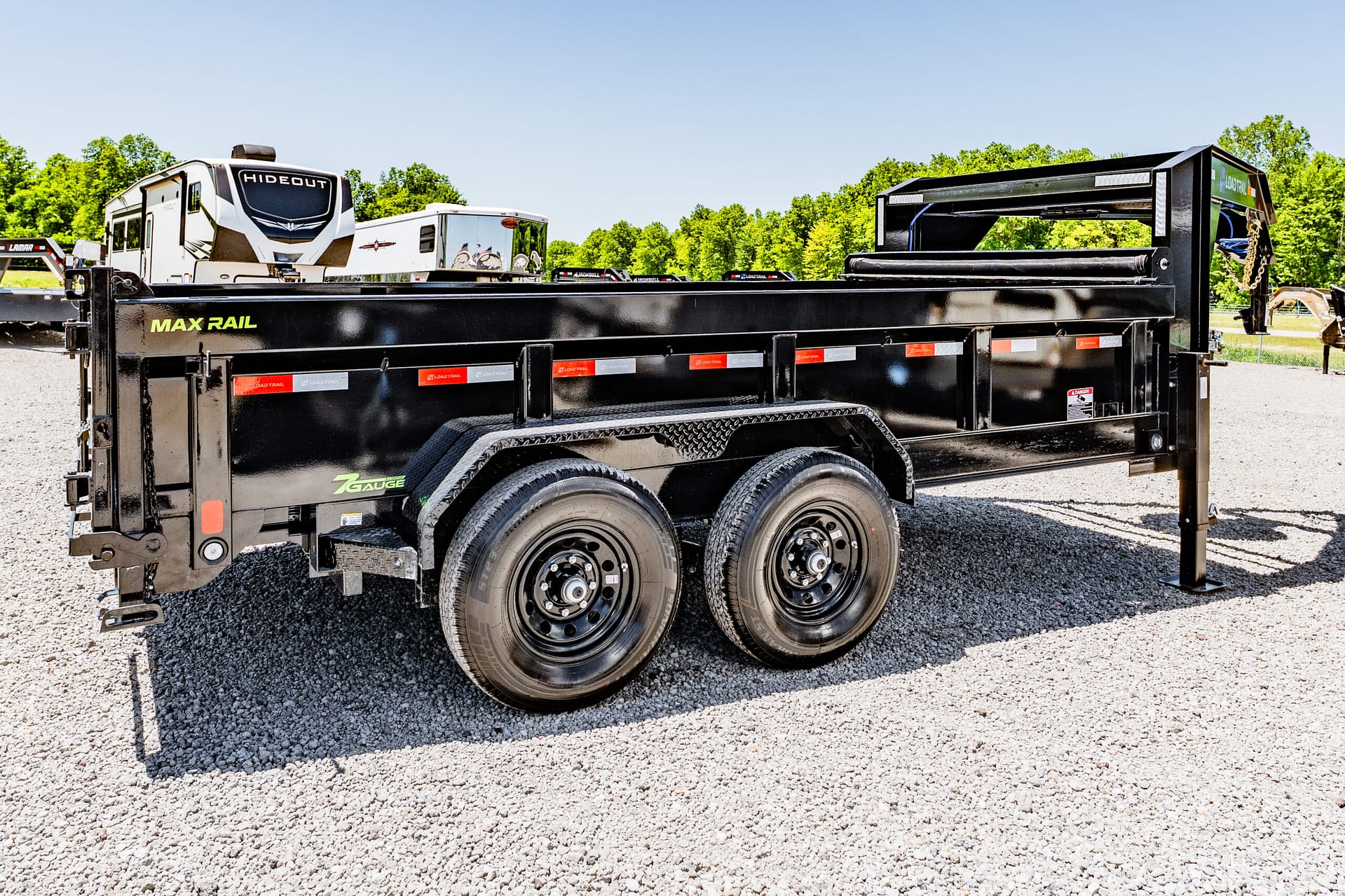 Black heavy-duty Max Rail flatbed trailer with dual axles on a gravel lot, trees in the background.