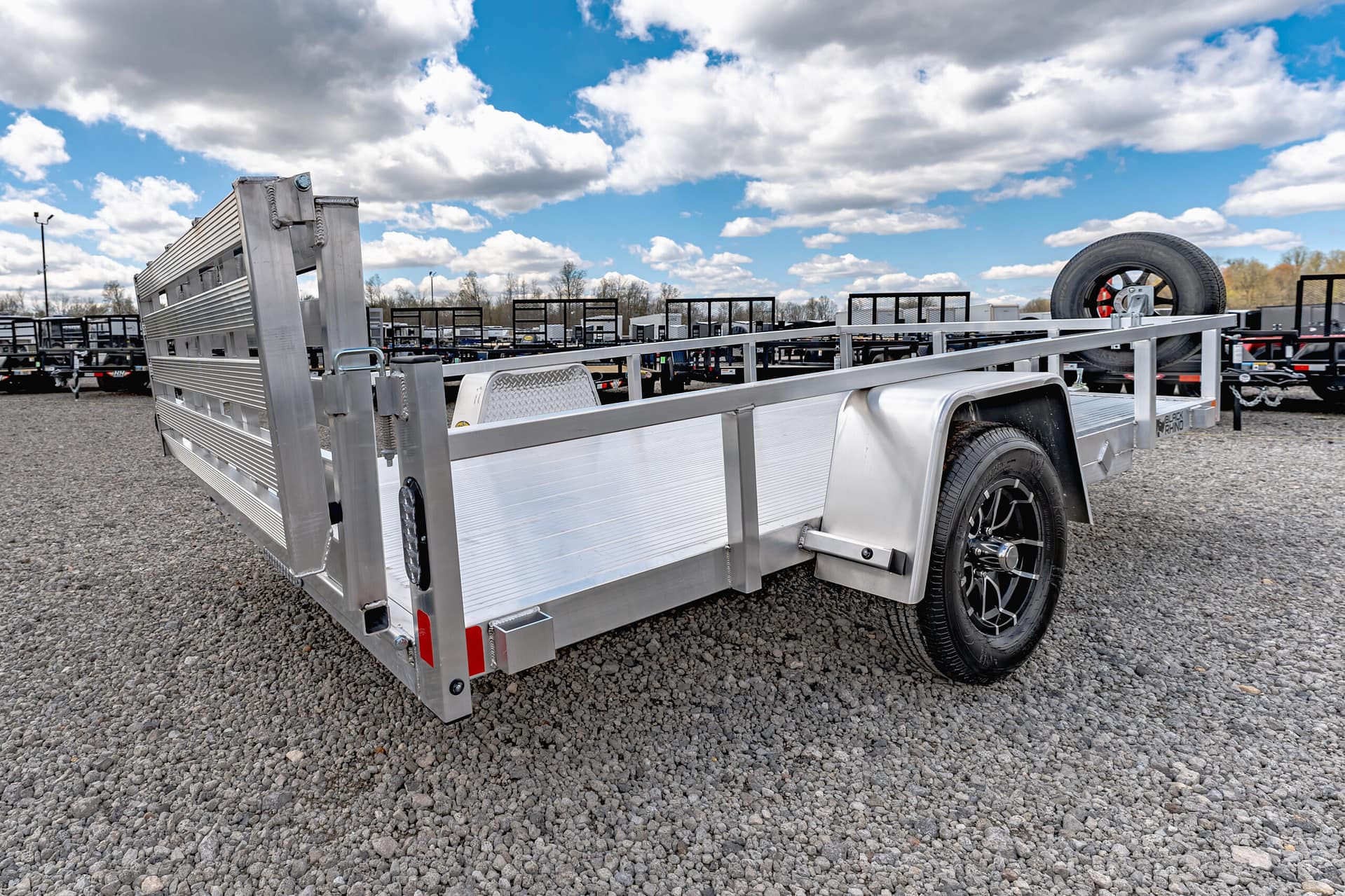 Aluminum flatbed trailer with a mounted spare tire at the rear, parked on a gravel lot with other trailers in the background under a blue sky with clouds.