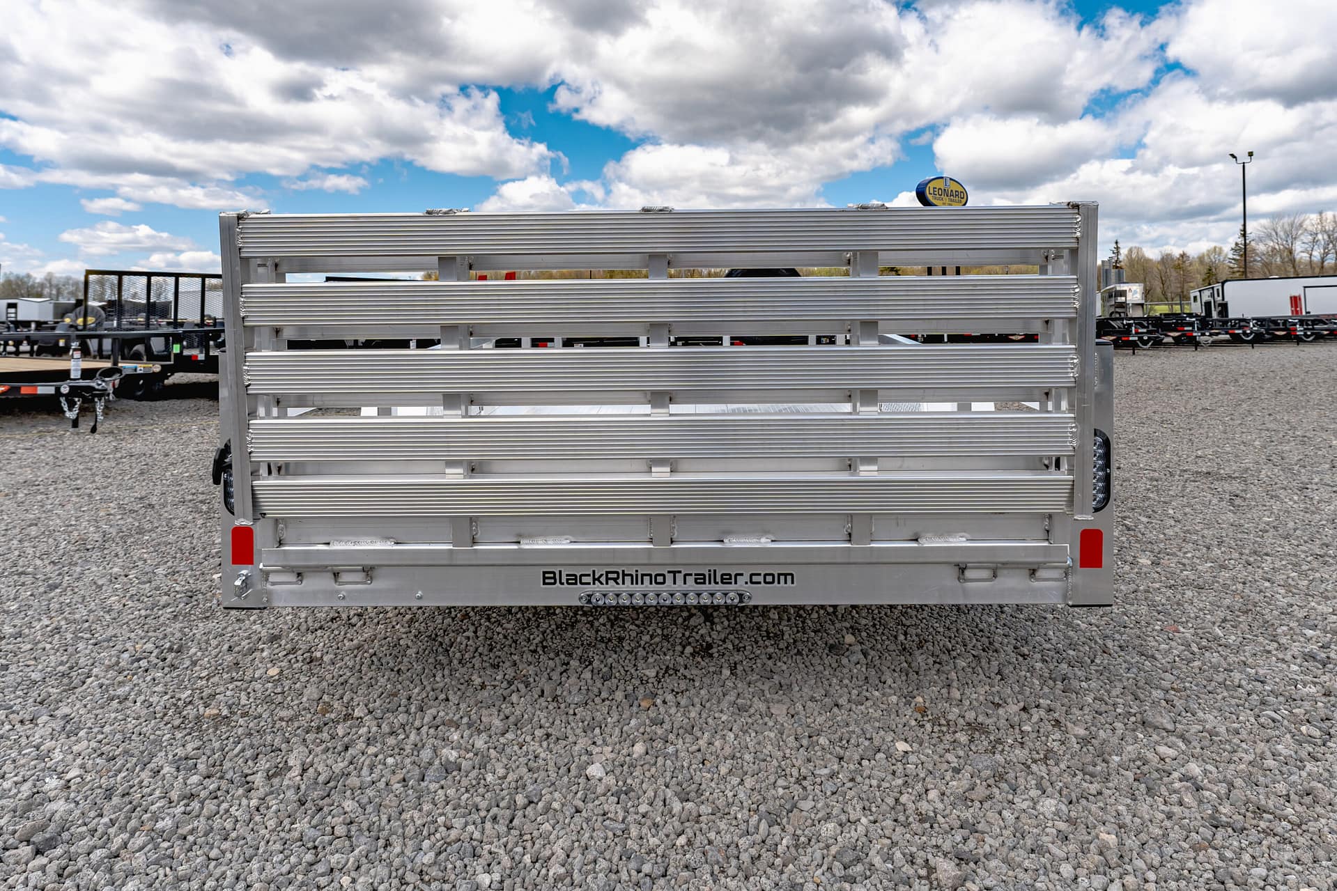Rear view of a silver aluminum trailer with 'BlackRhinoTrailer.com' branding, on a gravel lot with other trailers in the background.