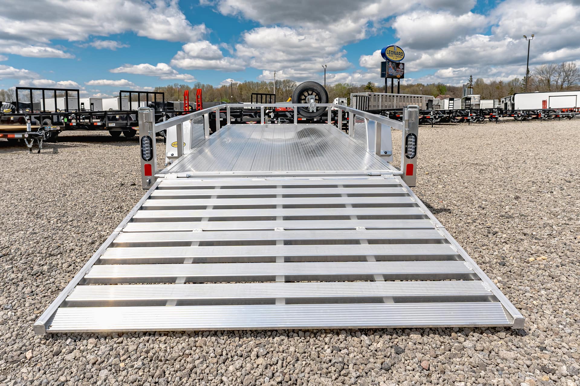 Low-angle view of a silver aluminum trailer ramp extending toward the camera in a gravel lot, with rows of trailers and a spare tire mounted on a hitch in the distance under a blue, cloudy sky at a trailer yard.