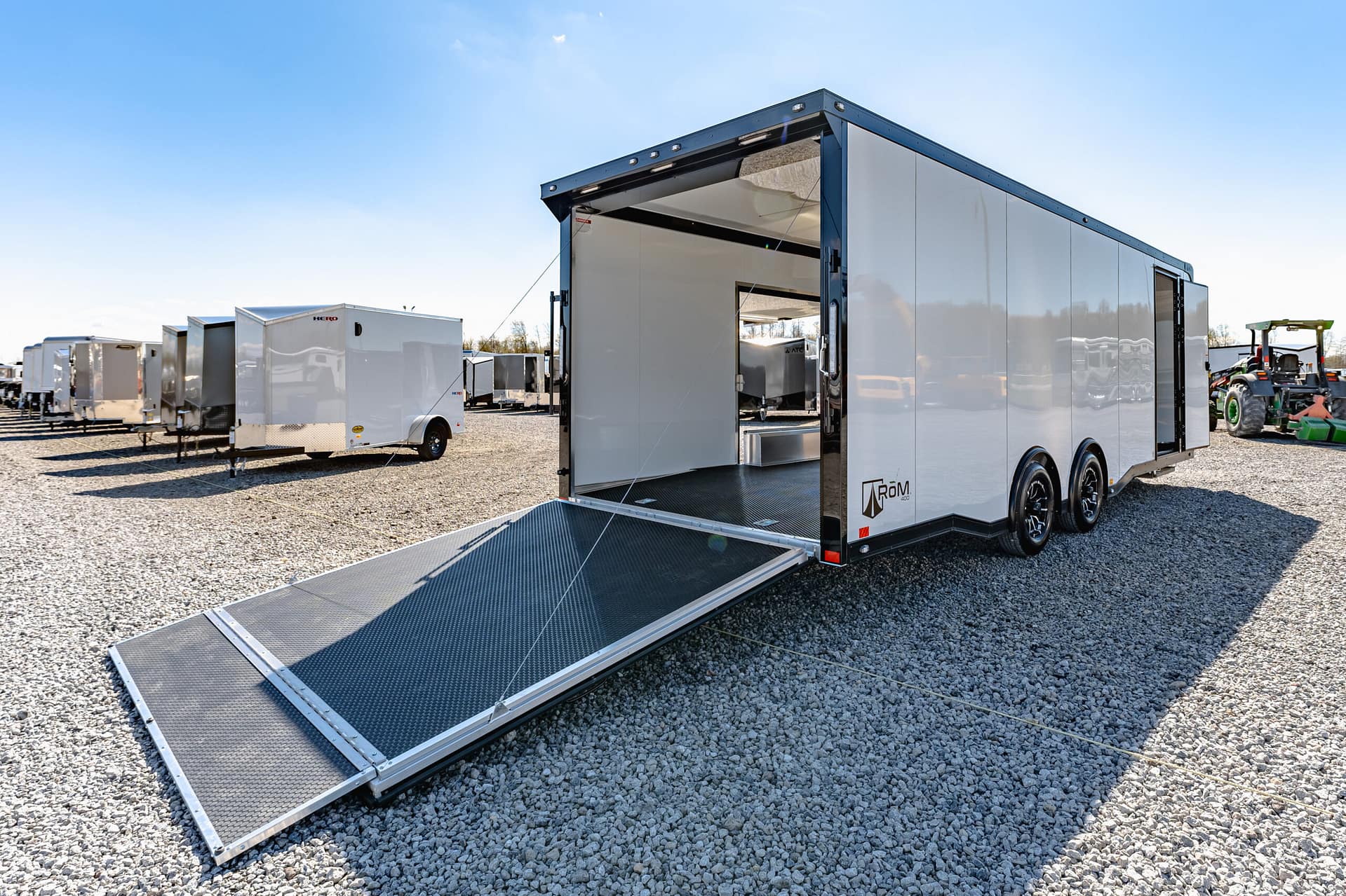 Open side of a white enclosed trailer with a metal loading ramp extended to the gravel ground, ready for loading, in a lot with other trailers under a clear blue sky.