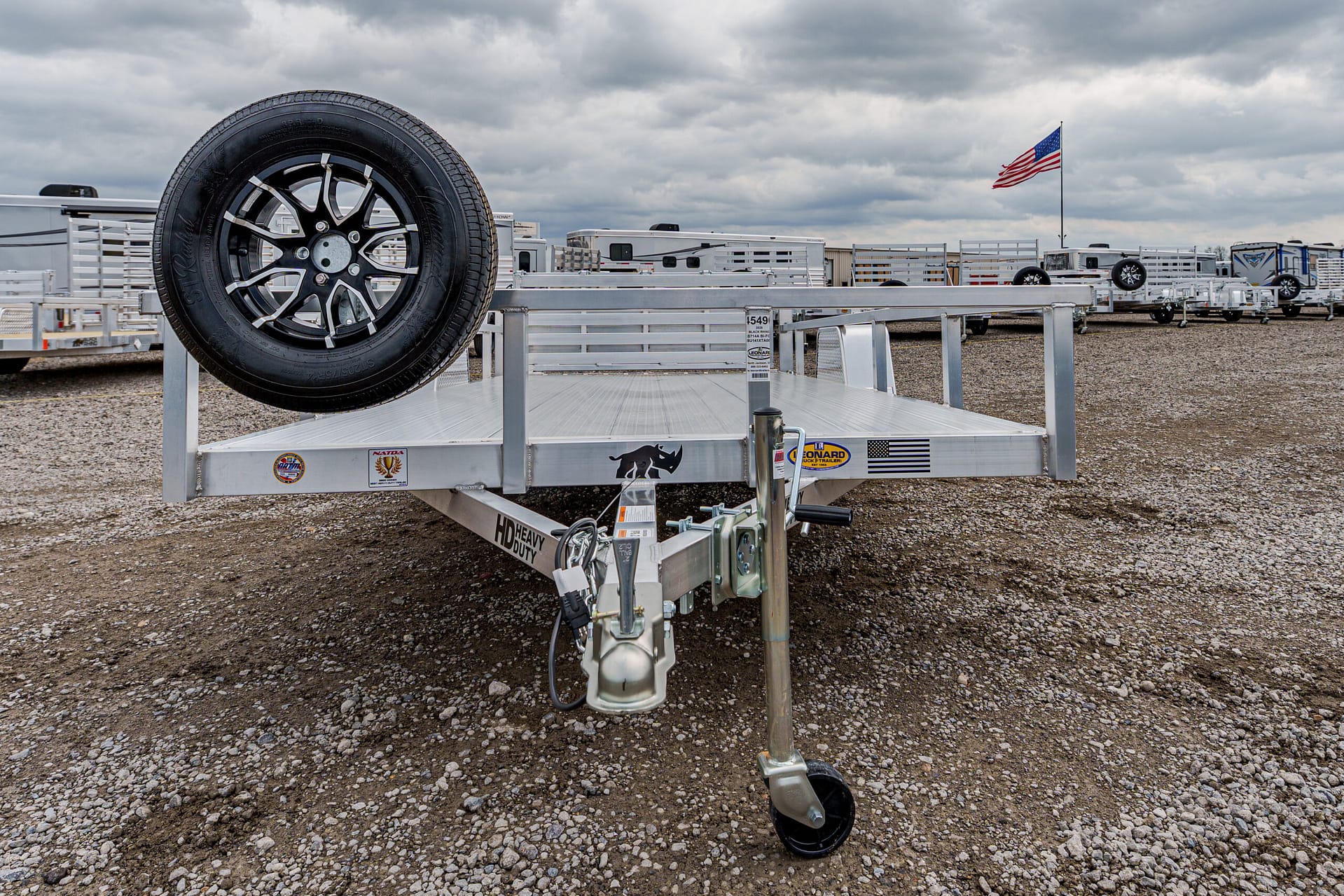 Aluminum flatbed trailer with a mounted spare tire on the left, hitch and jack at the front, gravel lot and other trailers in the background under a cloudy sky between a distant American flag and vehicles.