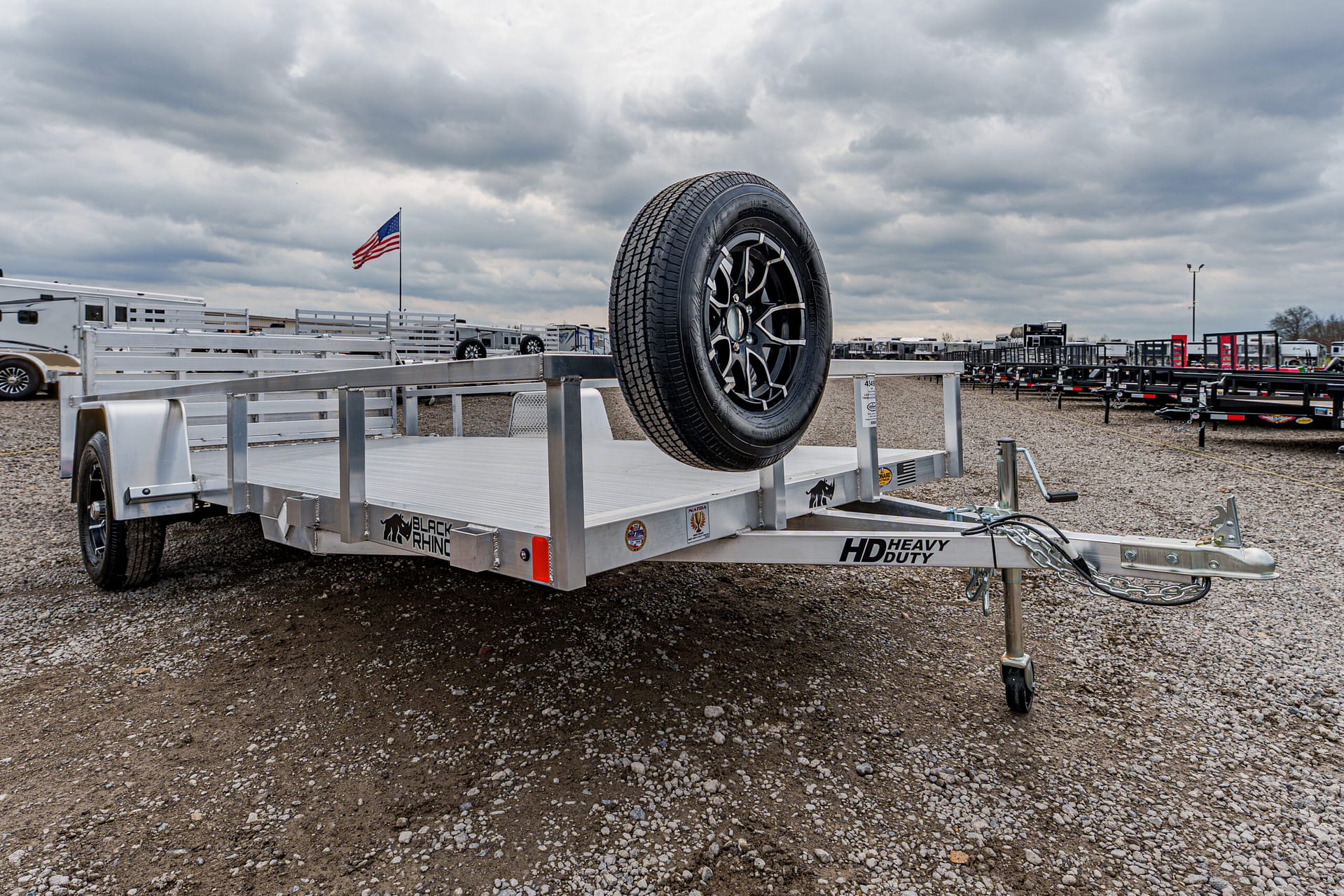 Aluminum flatbed trailer with a mounted spare tire on its front, parked on a gravel lot with other trailers and an American flag in the cloudy background.