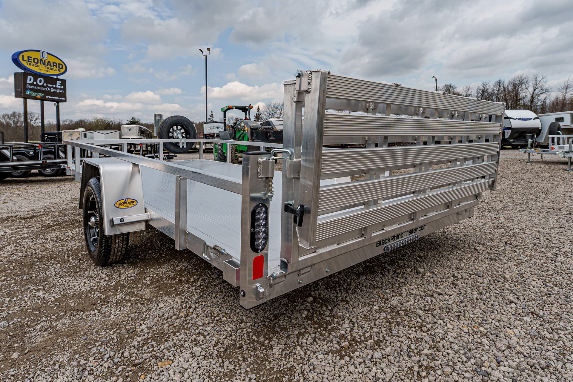 Aluminum utility trailer with a fold-down ramp gate on a gravel lot at a trailer dealership.