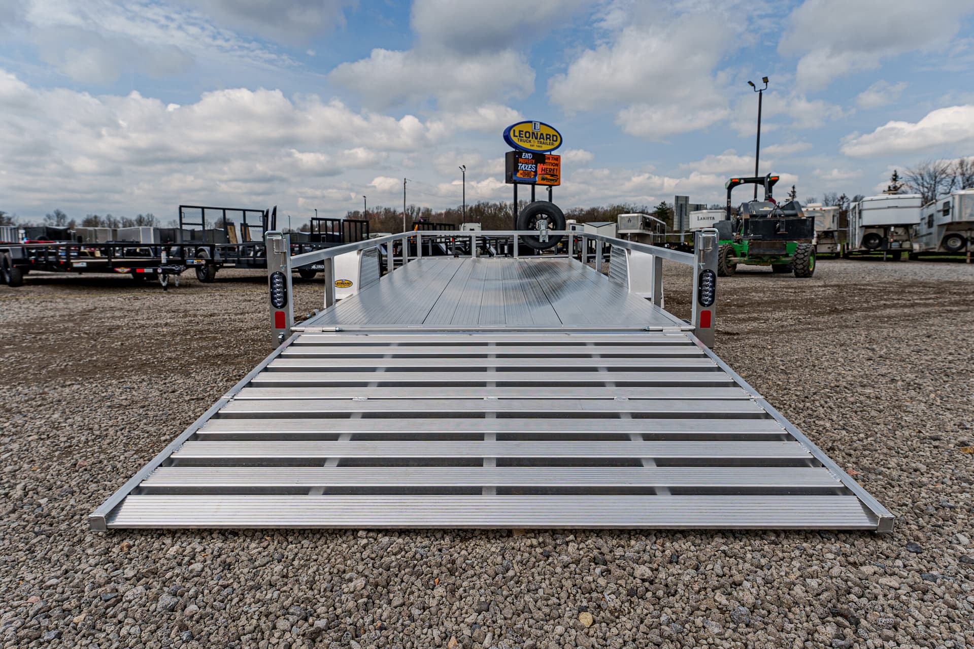 Silver aluminum car trailer with a lowered ramp, parked on a gravel lot with other trailers in the background.