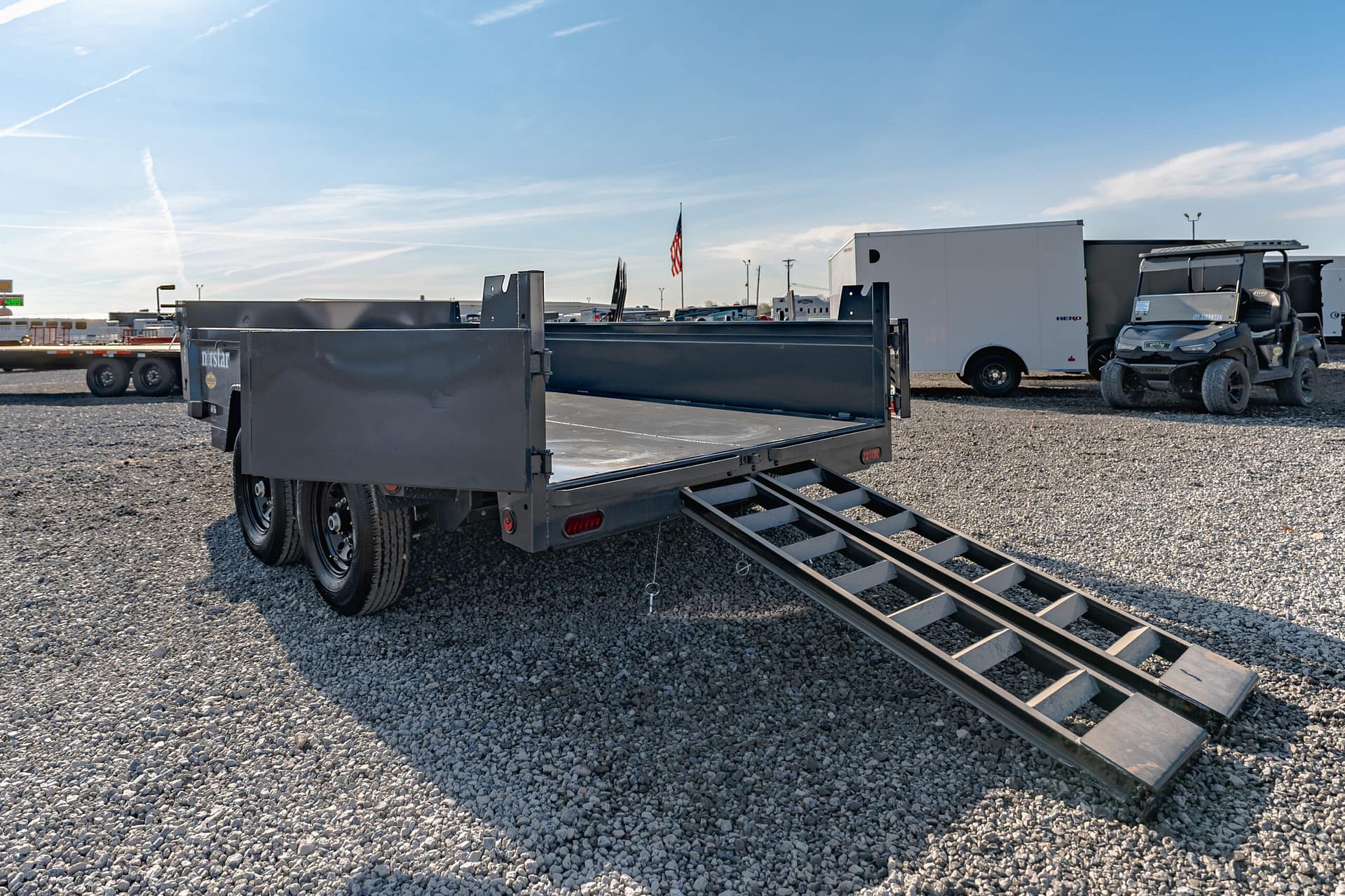 Gray flatbed trailer with folded ramps on a gravel lot, a small utility vehicle on the right, and a white service trailer in the background with an American flag nearby.