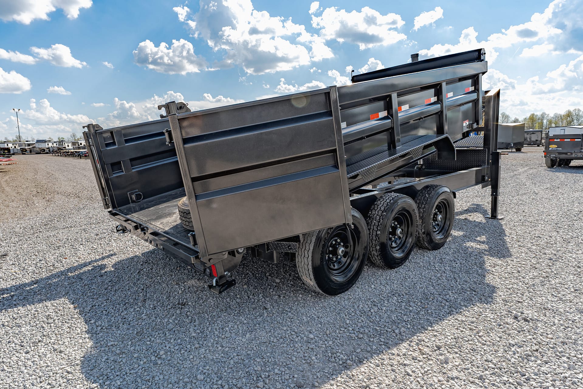 Black dump trailer tilted open on a gravel lot, with other trailers in the background under a blue, partly cloudy sky.