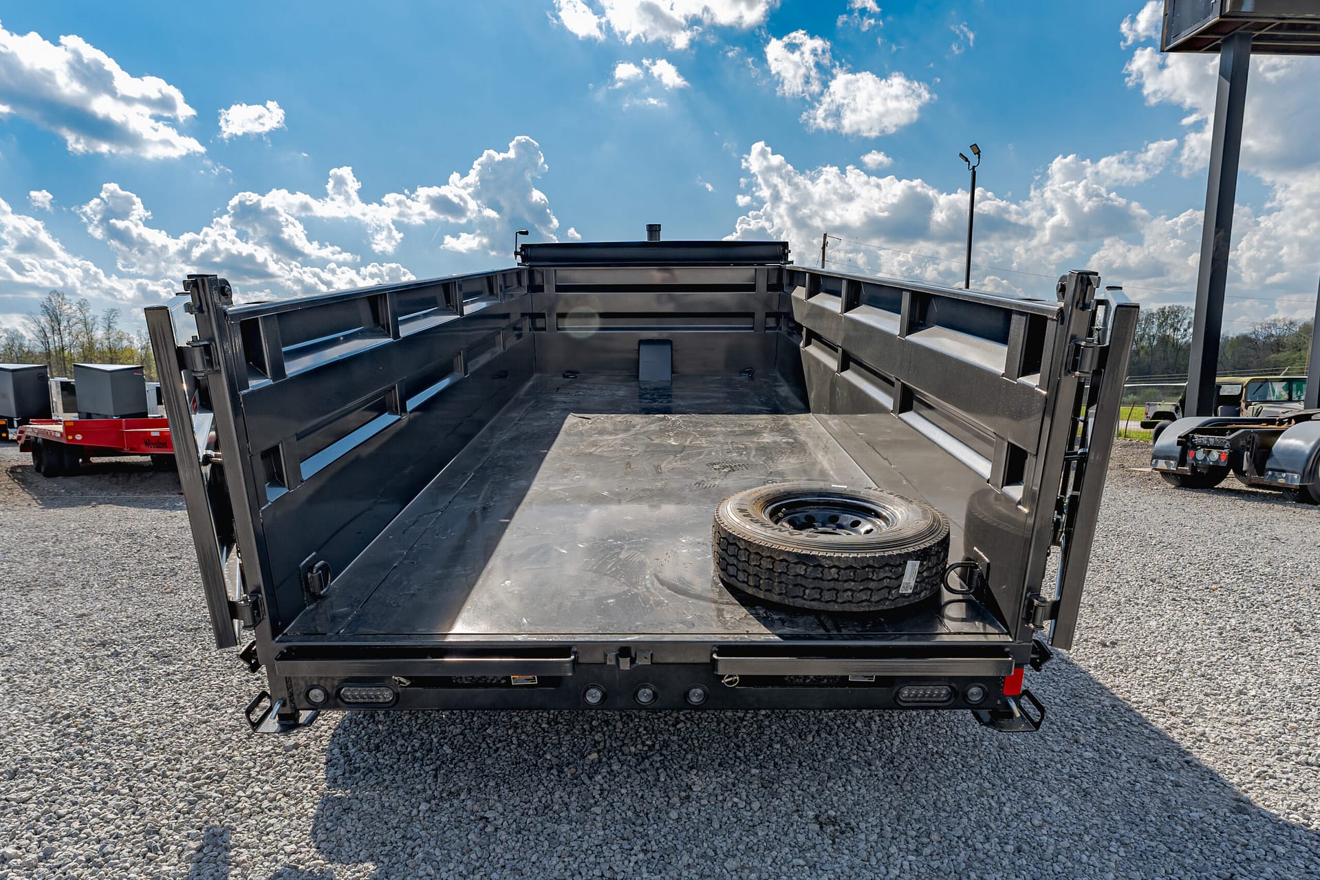 Open black dump trailer bed with a spare tire inside on a gravel lot under a blue sky.